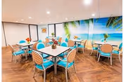 Dining area with several tables and turquoise chairs in front of a large tropical beach mural on the wall.