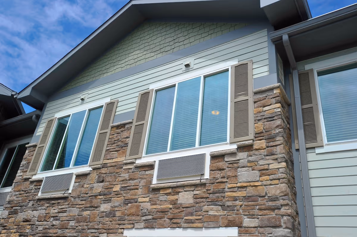 Exterior view of a building wall with stone and siding finishes, featuring three large windows with beige shutters and air conditioning units below two of the windows, under a blue sky with some clouds.