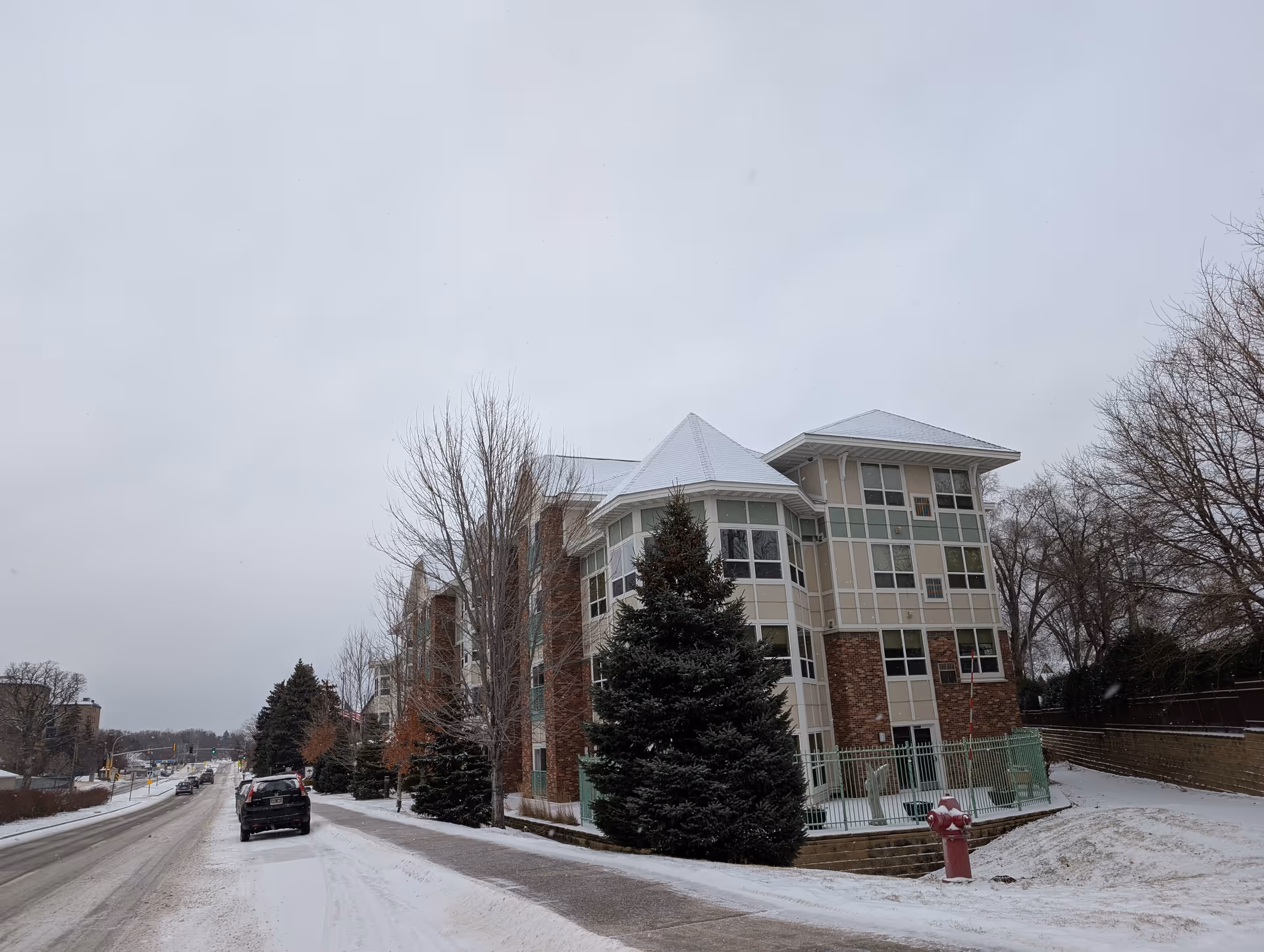 Multi-story senior living building beside a snowy street with parked cars and evergreen trees.