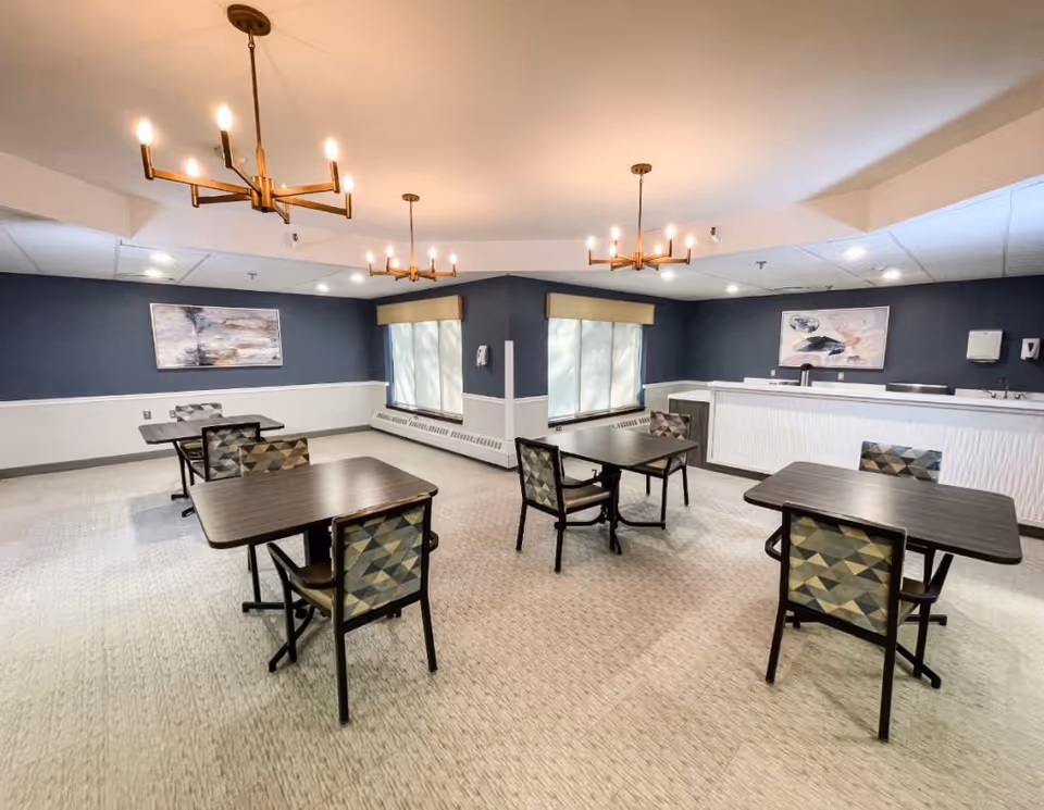 A dining room with four dark wood tables, each paired with two chairs featuring geometric patterned upholstery. The room has blue walls with white wainscoting, two large windows with beige valances, and two abstract paintings on the walls. Three modern chandeliers with exposed bulbs hang from the ceiling, and there is a white counter area along one wall.