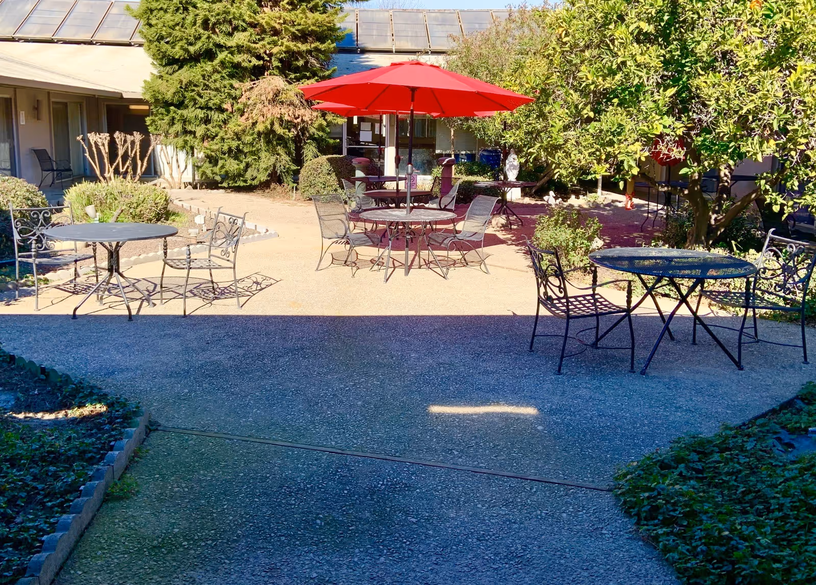 Outdoor courtyard area with metal tables and chairs, some shaded by a large red umbrella. Surrounding the courtyard are bushes, trees, and a building with solar panels on the roof.