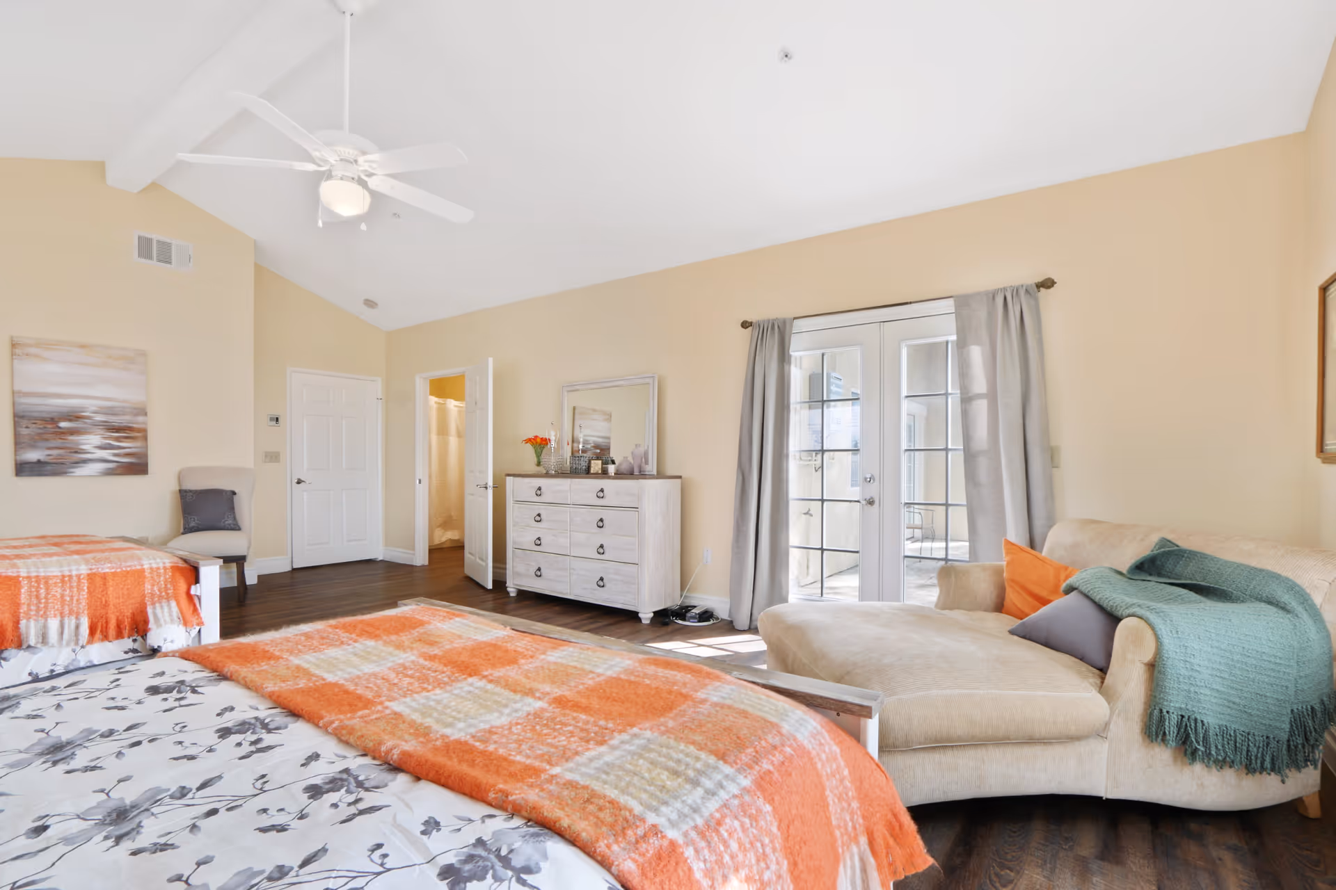A bright and spacious bedroom with two beds covered in floral and orange plaid blankets. The room features a beige chaise lounge with colorful pillows and a green throw, a dresser with a mirror and flowers on top, a chair, and French doors with gray curtains leading to an outdoor area. The walls are painted light beige and the floor is dark wood.