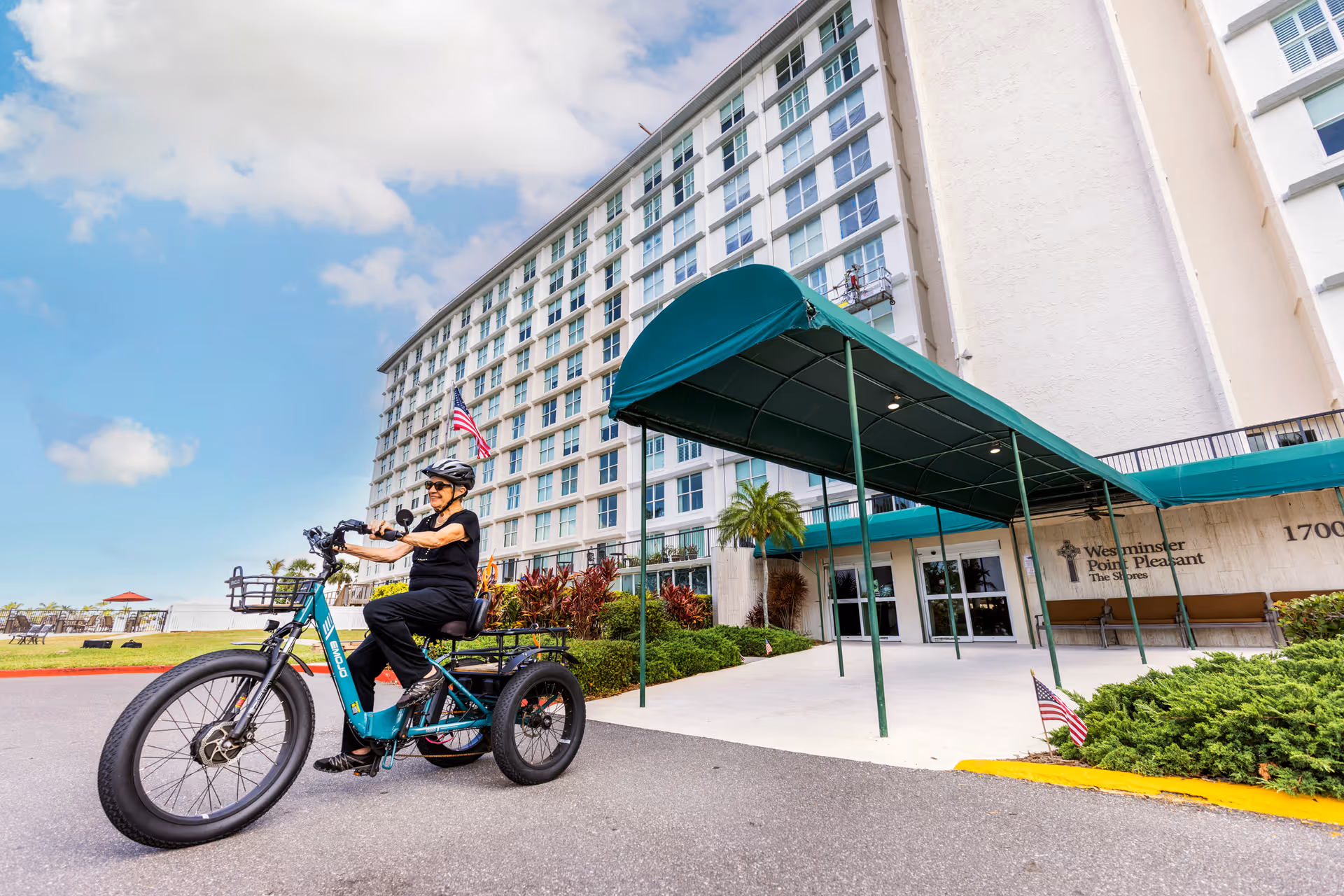 A senior woman wearing a helmet rides a teal three-wheeled electric bike outside the entrance of Westminster Bradenton - Point Pleasant Neighborhood, a multi-story senior living facility. The building has many windows and a green awning over the entrance. The sky is partly cloudy and there are some plants and an American flag near the entrance.