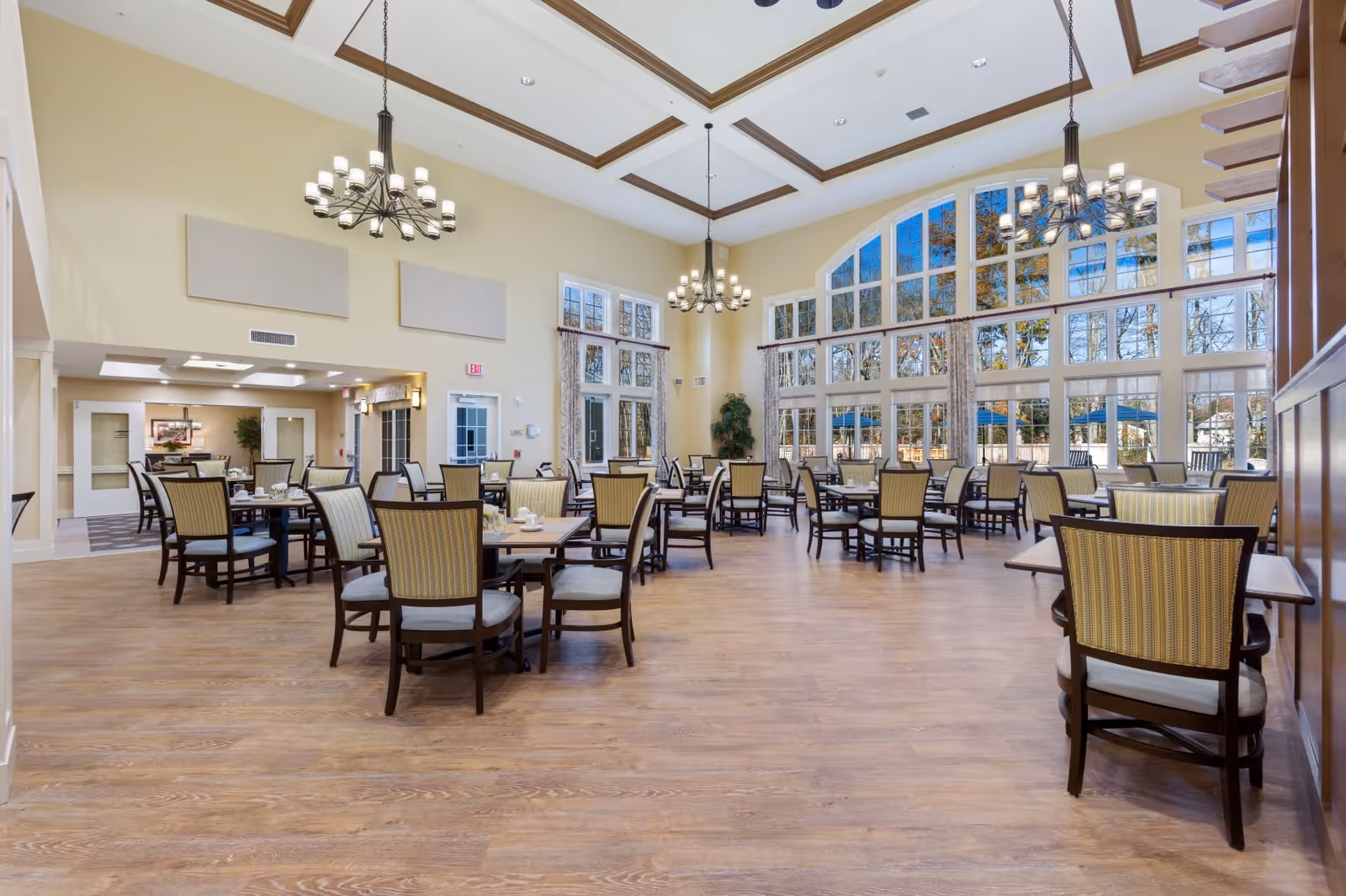 Spacious dining room with multiple tables and chairs arranged neatly on a wooden floor. Large windows with an arched design allow natural light to fill the room, and two chandeliers hang from the high ceiling. The room has light-colored walls and a calm, inviting atmosphere.