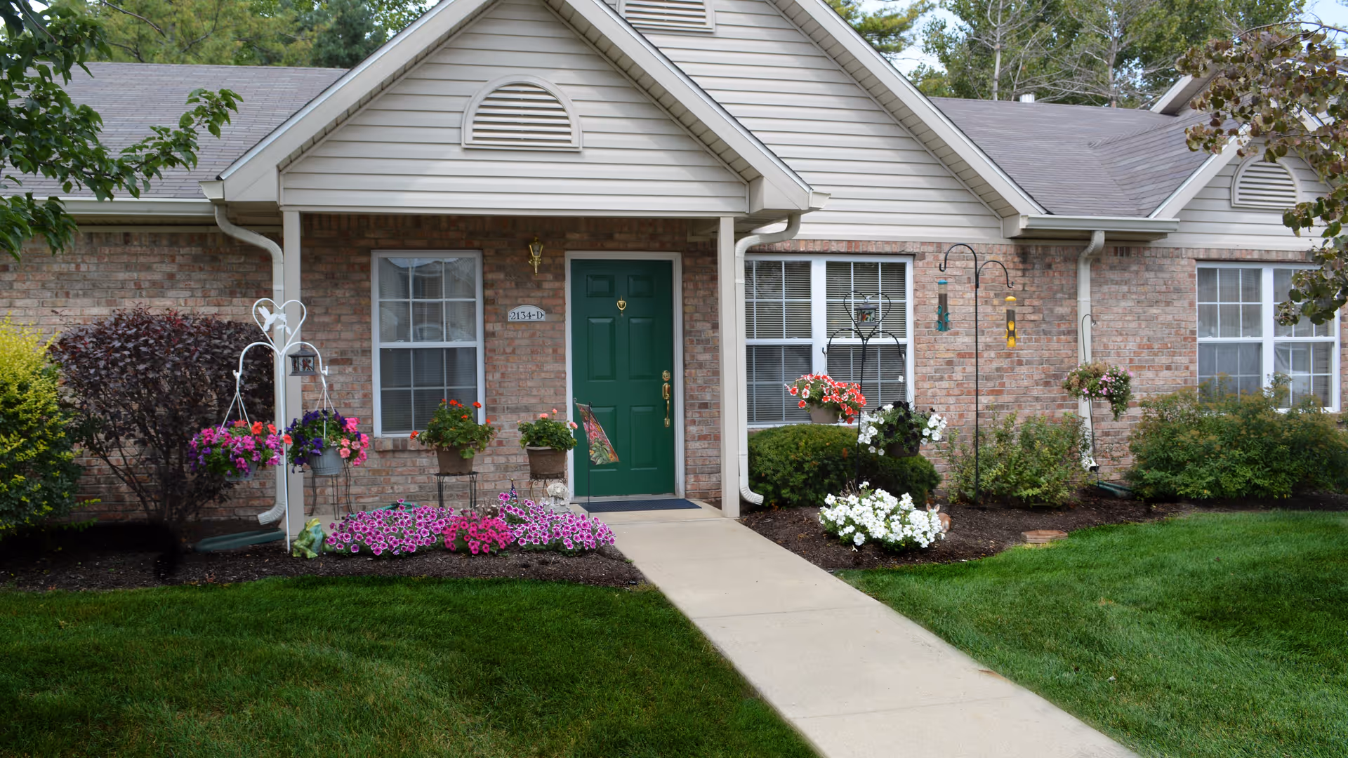 Front entrance of a single-story brick unit with a green door, concrete walkway, and landscaped flower beds.