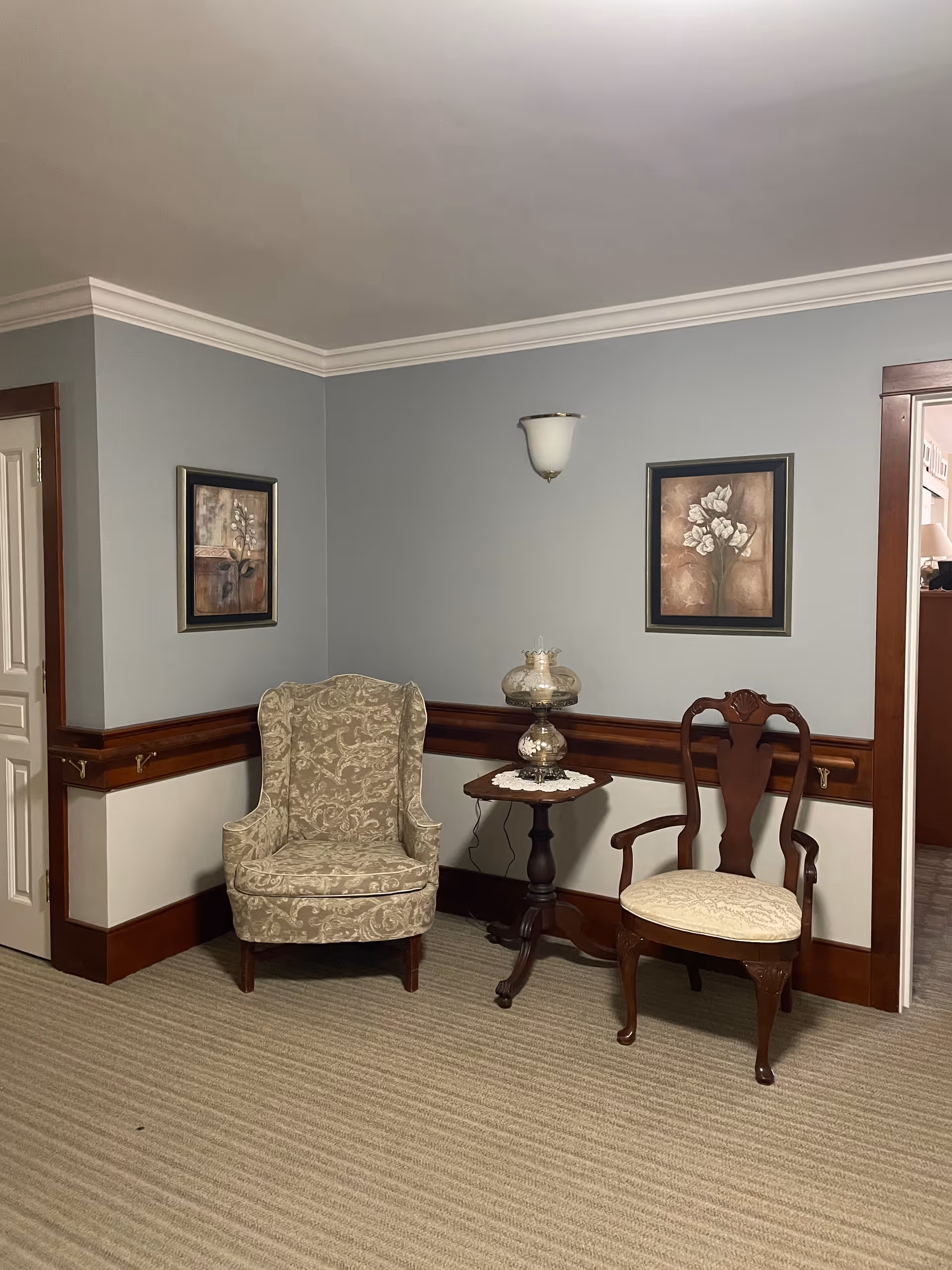 A corner of a room with two chairs, one upholstered armchair with a floral pattern and one wooden chair with a cushioned seat. Between the chairs is a small wooden side table with a decorative lamp on a doily. The walls are painted light gray with wooden trim and two framed floral paintings hanging above the chairs. The floor is carpeted with a beige striped pattern.