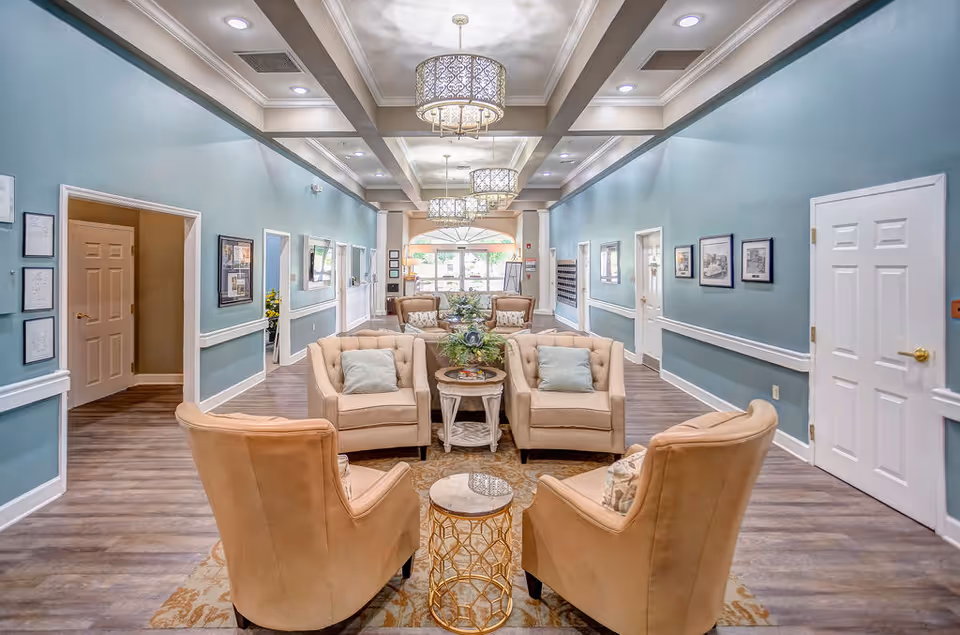 A spacious and well-lit common area with light blue walls and wood flooring. The room features multiple beige armchairs arranged around small tables with decorative plants. The ceiling has recessed lighting and elegant hanging light fixtures. Several doors and framed pictures line the walls, and large windows at the far end let in natural light.