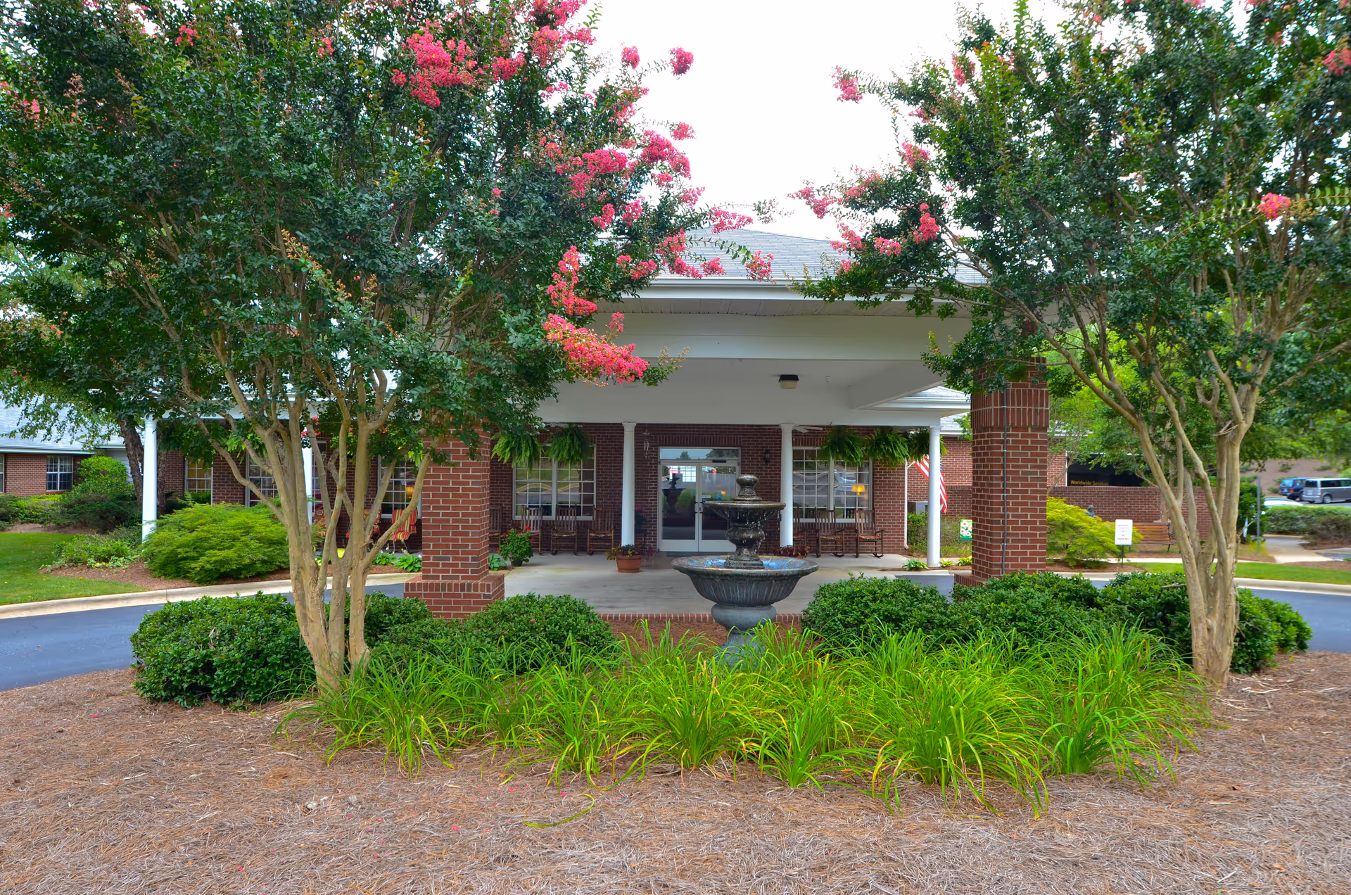 Front entrance of a brick building with a covered driveway supported by white columns. There is a tiered water fountain in the center of a landscaped area with green bushes and flowering trees with pink blossoms. The building has large windows and rocking chairs on the porch.