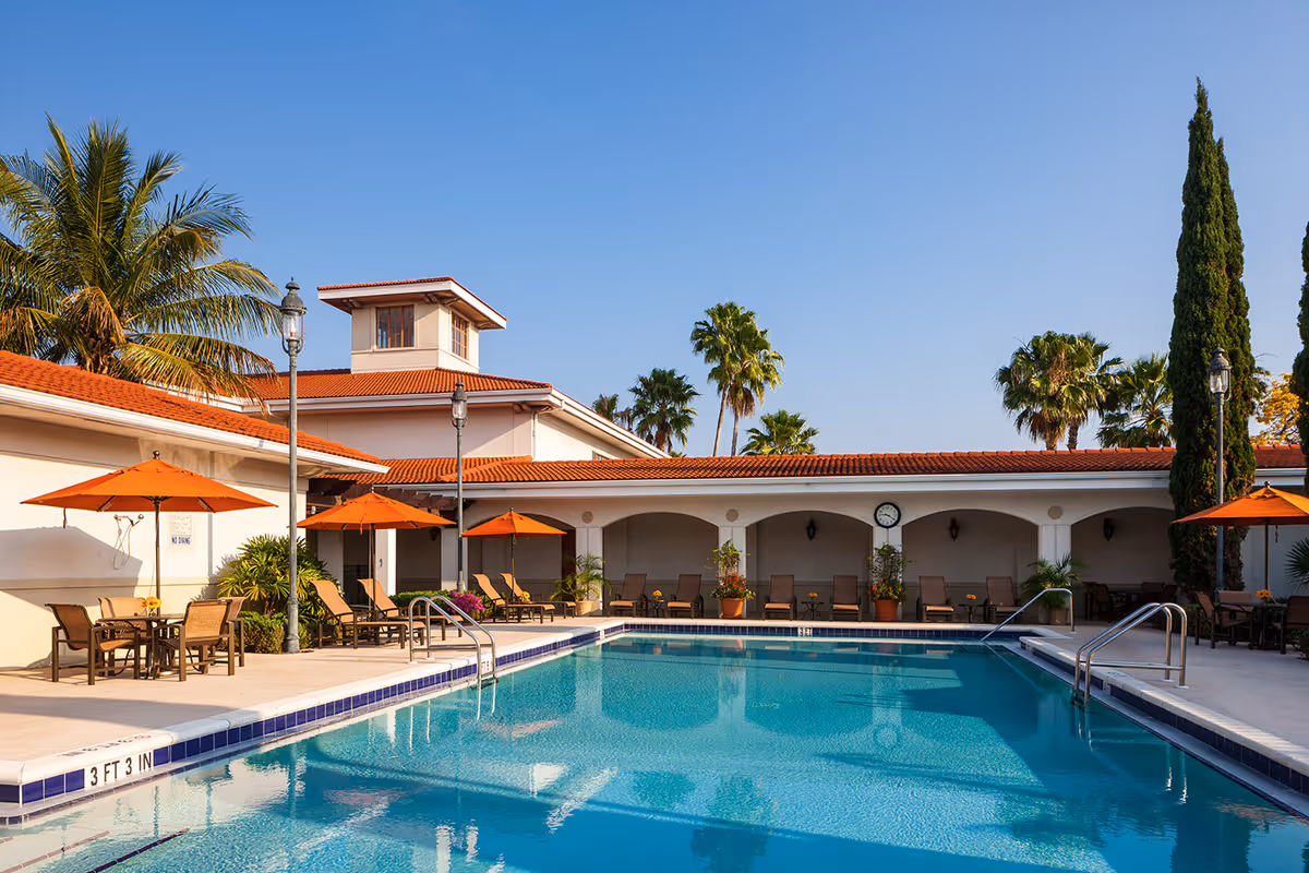 Outdoor swimming pool with lounge chairs, orange umbrellas, and a Mediterranean-style building in the background.