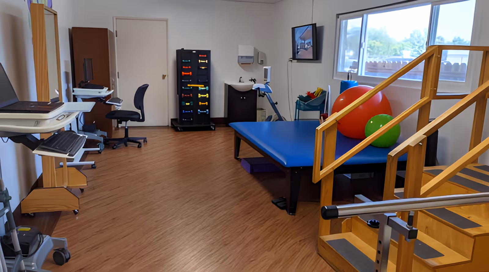 A physical therapy room with wooden stairs and handrails for rehabilitation exercises, a blue padded therapy table with large red and green exercise balls on it, a rack of colorful dumbbells, a sink, a chair, and a computer workstation on the left side. A window lets in natural light.