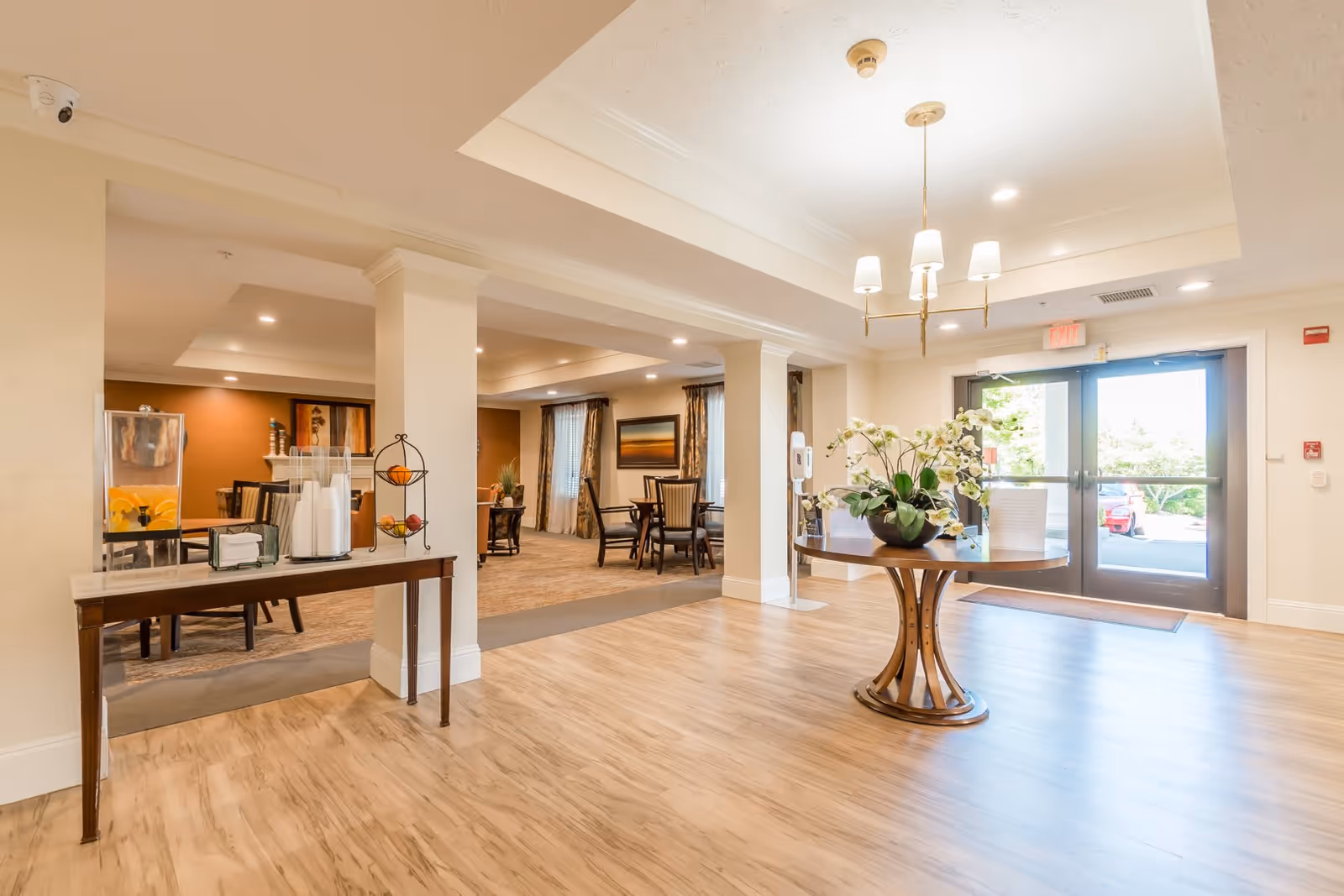 Bright and spacious interior of a senior living facility with wooden flooring, a round table with a flower arrangement in the center, and a side table with a beverage dispenser and cups. There are multiple seating areas with tables and chairs in the background, and large glass doors letting in natural light.