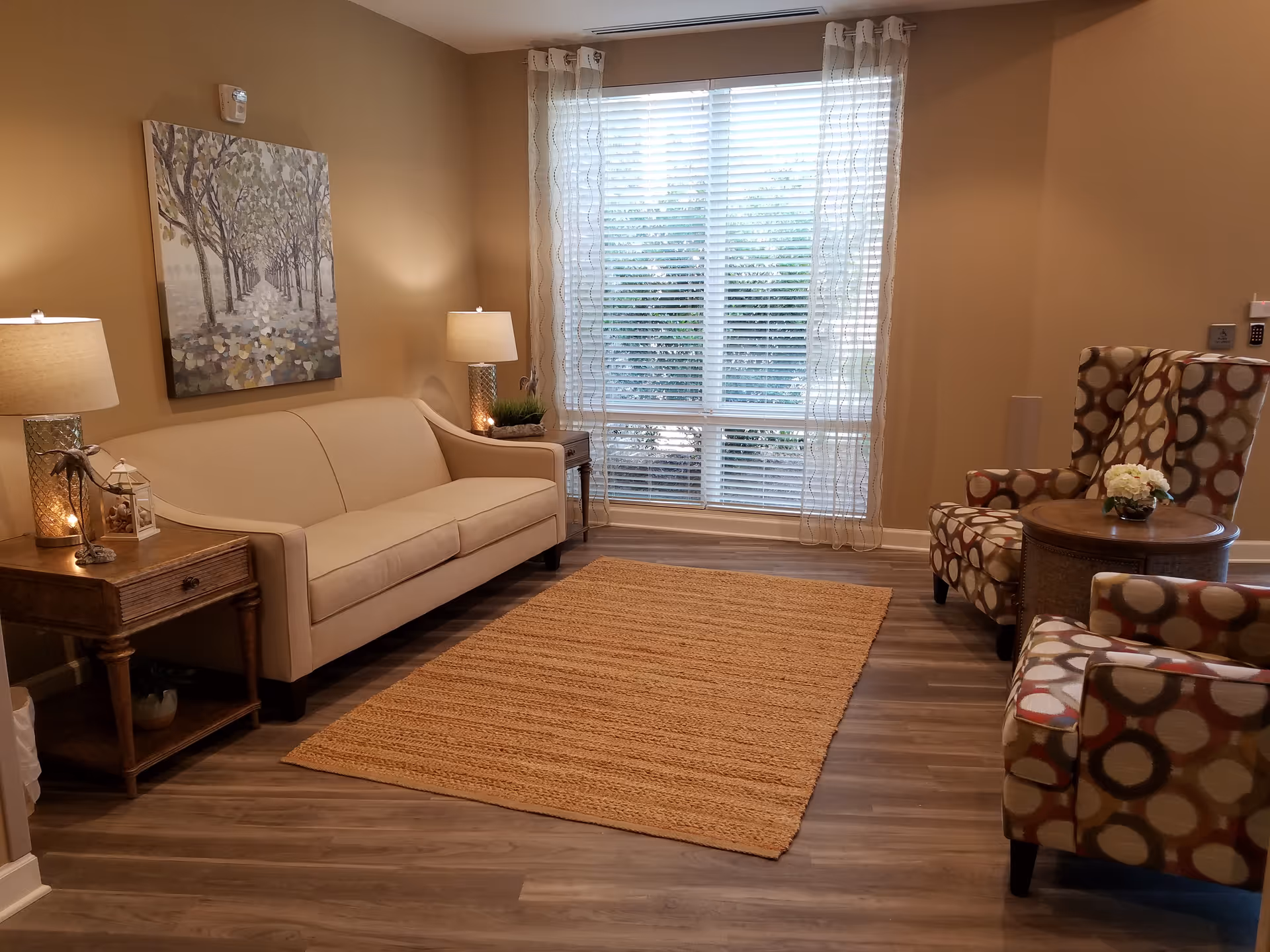 A cozy living room with a beige sofa, two patterned armchairs, a wooden side table with lamps, a round wooden coffee table with a flower arrangement, a large window with sheer curtains, and a painting of a tree-lined path on the wall.