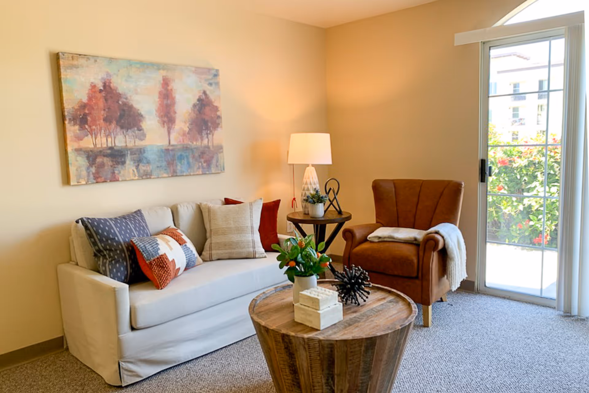 A bright living room with a white sofa, brown armchair, wooden coffee table, wall artwork, and a sliding glass door to the outside.