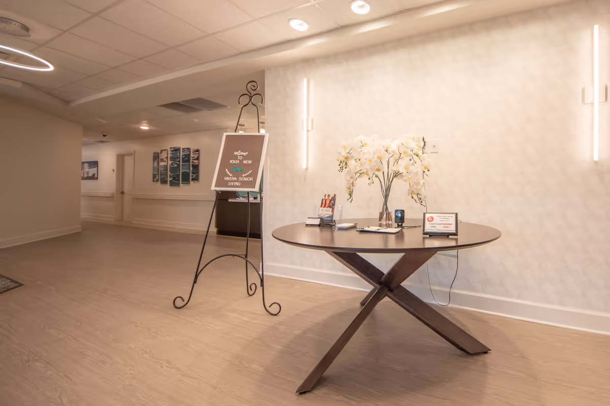 Well-lit senior living facility lobby with a round table displaying brochures and a vase of flowers and a welcome sign on an easel.
