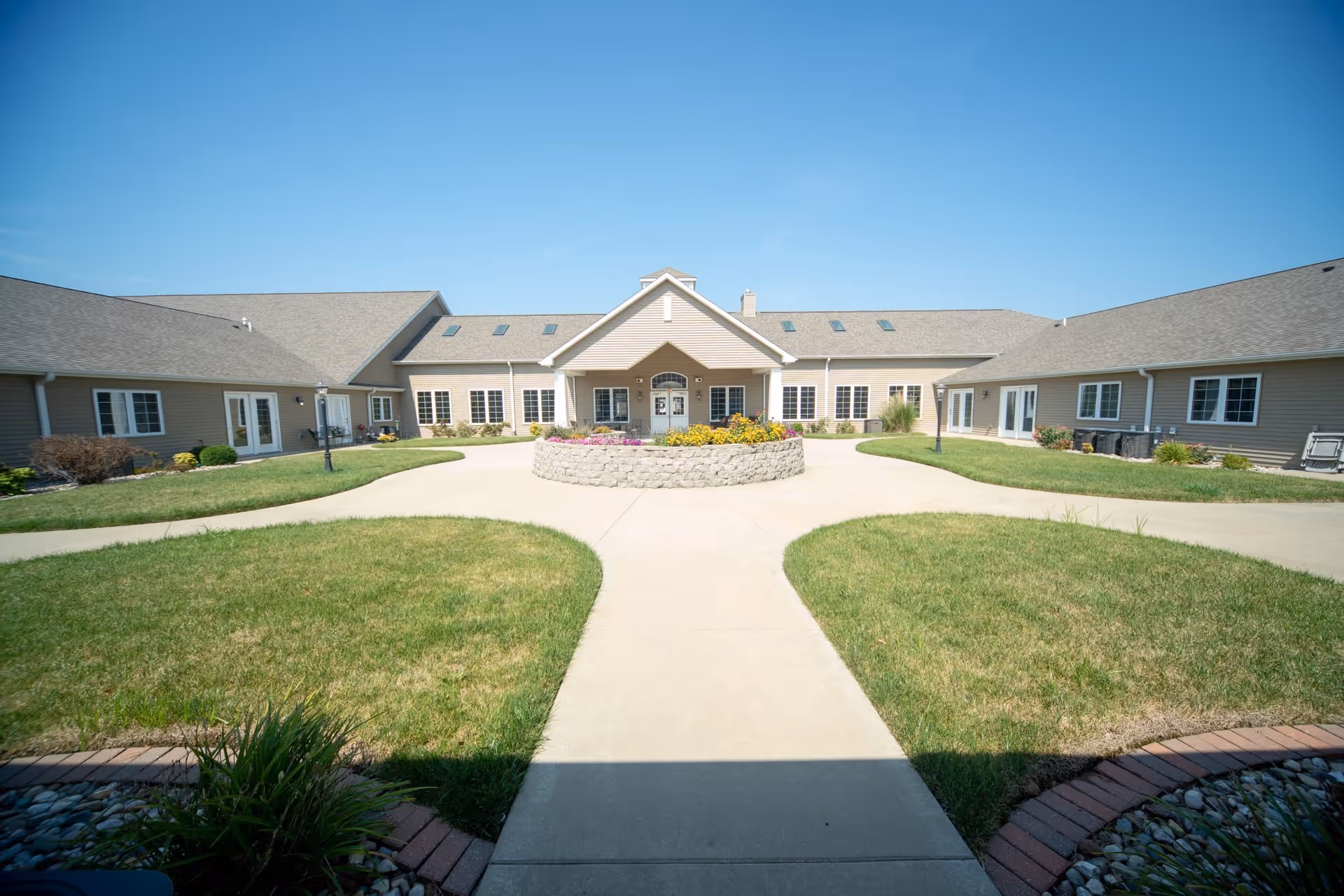 Exterior view of Villas of Holly Brook Herrin showing a single-story building with beige siding and multiple windows surrounding a circular driveway with a central flower bed under a clear blue sky.