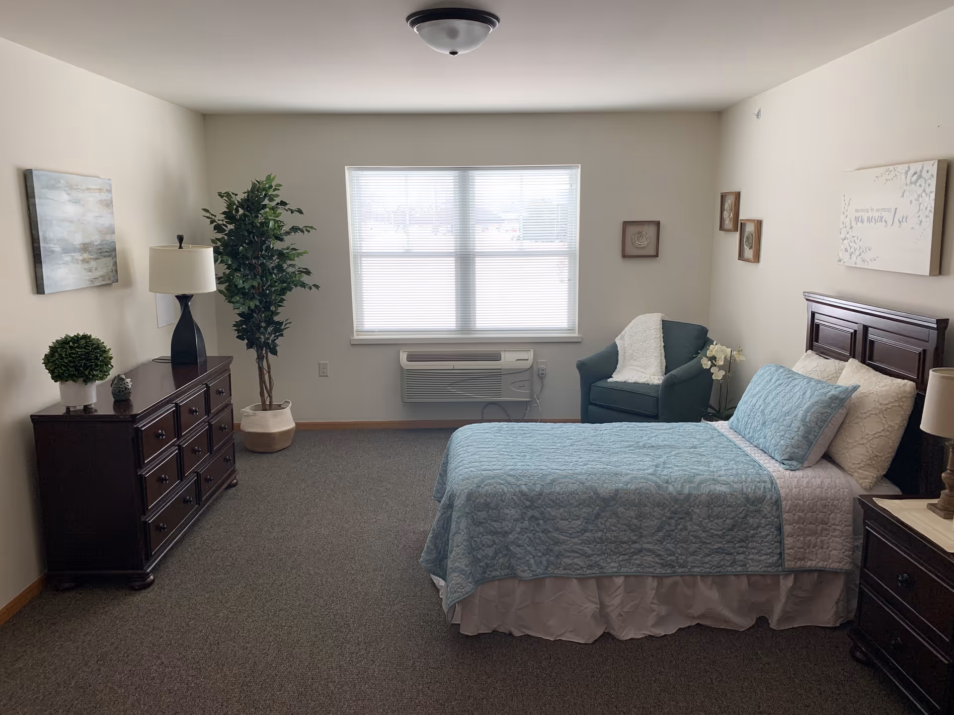 A neatly arranged bedroom with a single bed covered in a light blue quilt and white pillows. There is a dark wooden dresser with a lamp and small plants on it, a green armchair with a white throw blanket, and a large window with blinds letting in natural light. The walls are decorated with framed artwork and a sign above the bed.