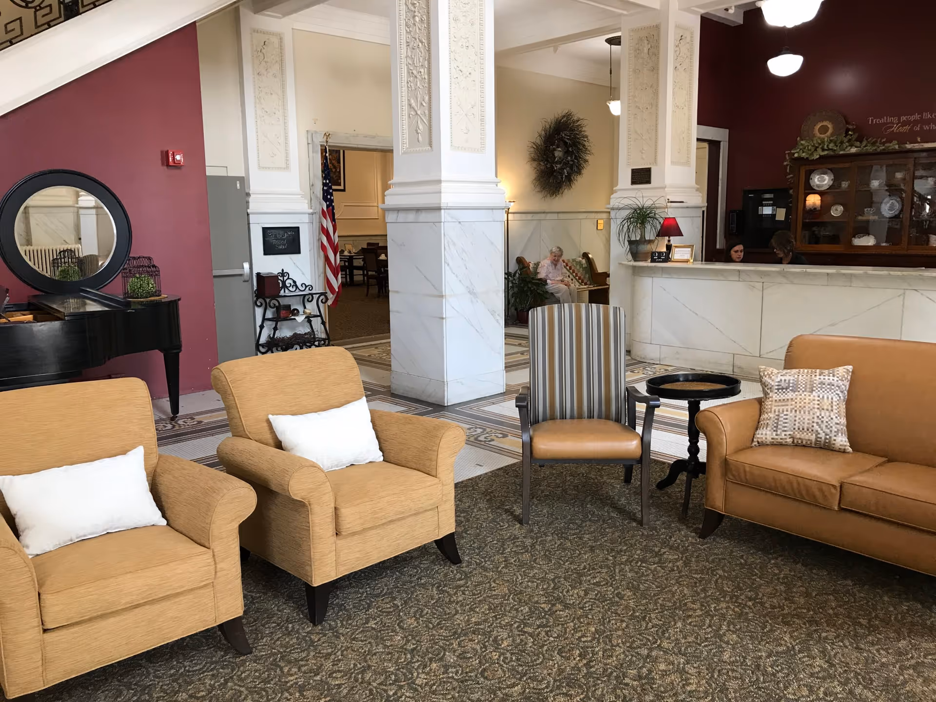 Lobby seating area with upholstered chairs and a sofa facing a reception desk and decorative marble columns.