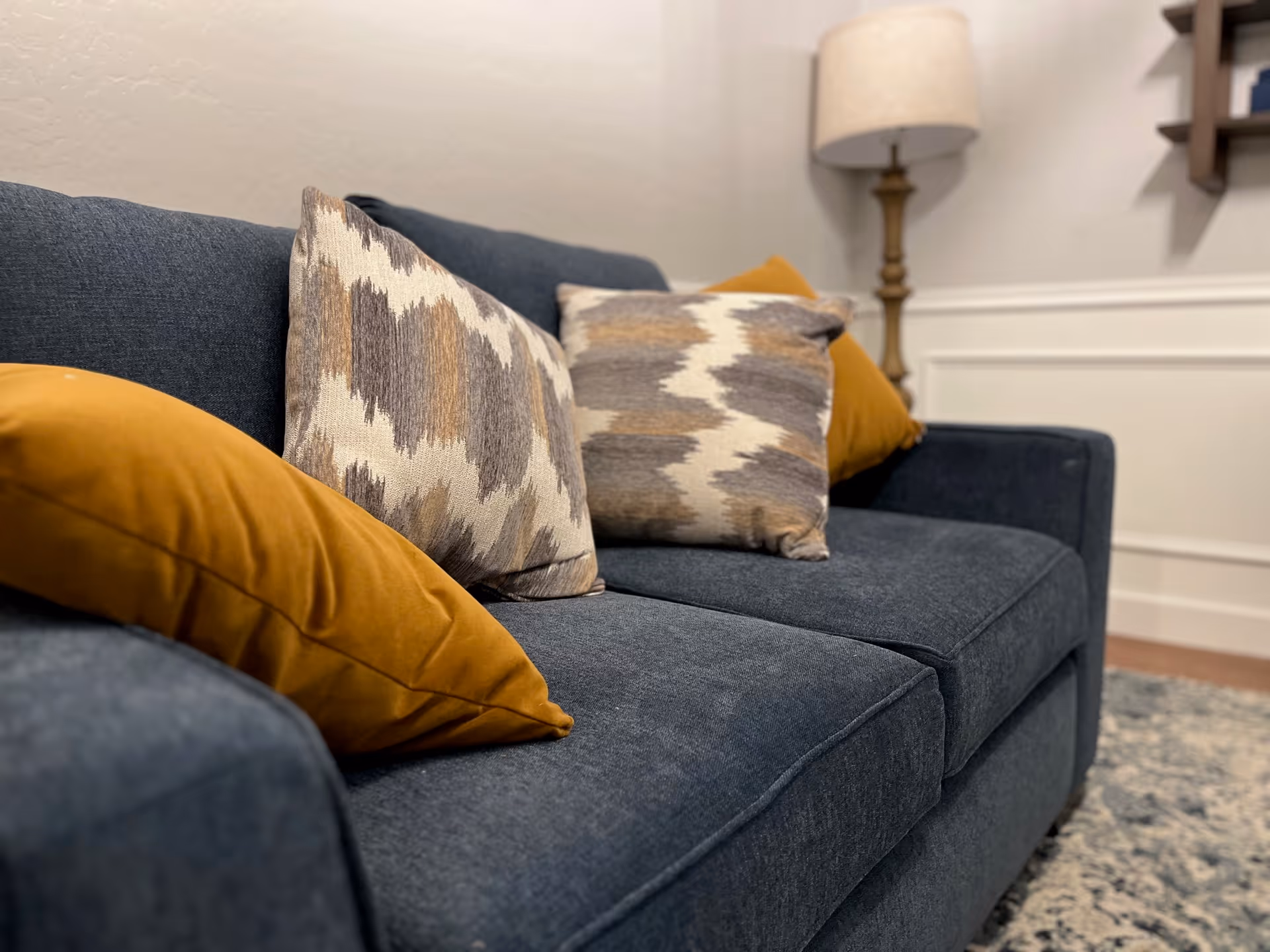 Blue upholstered sofa with patterned and mustard throw pillows, a lamp, and a rug in a living room setting.