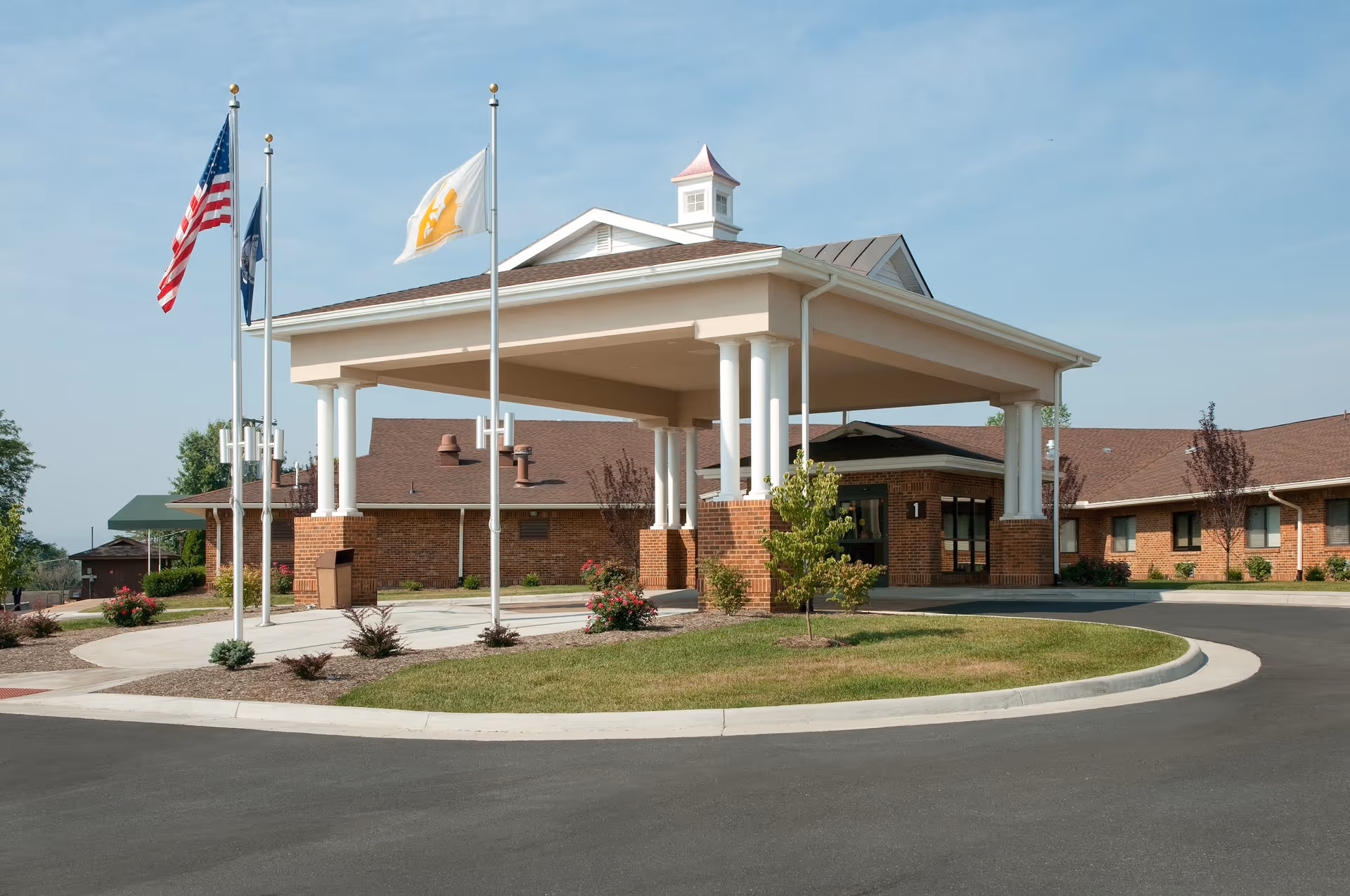 Front entrance of a single-story brick senior living facility with a covered drop-off portico and three flagpoles.