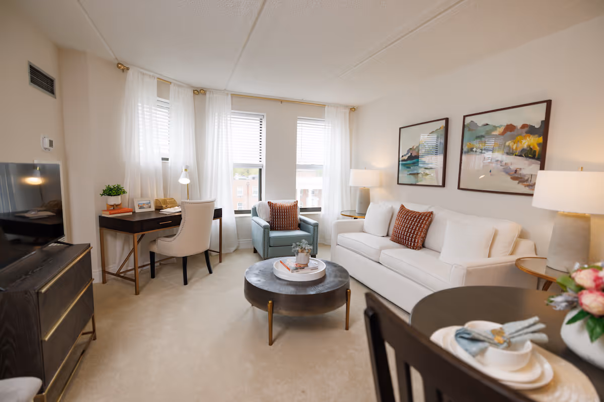 A bright and cozy living room with a white sofa adorned with patterned cushions, a blue armchair, a round coffee table with decorative items, and a small desk with a chair near two windows with white curtains. The room also features two framed landscape paintings on the wall, a TV on a dark wooden stand, and a round dining table with place settings and a flower arrangement.