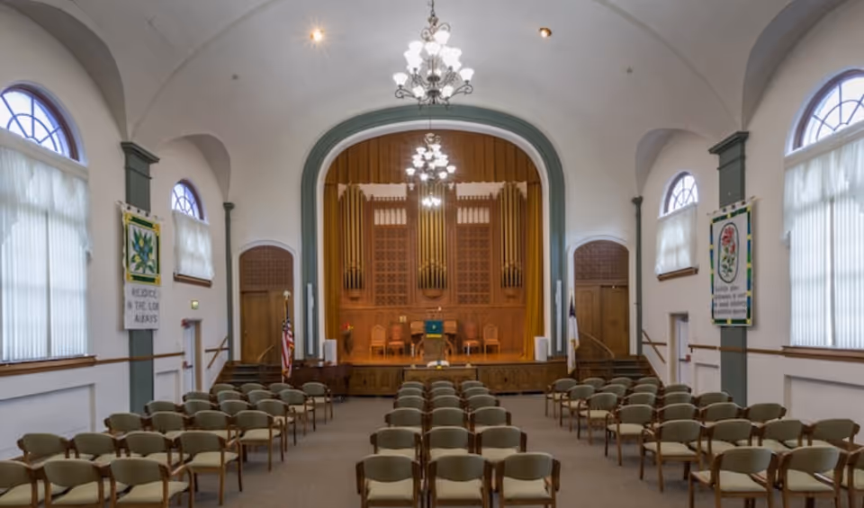 Interior view of a chapel or auditorium with rows of green cushioned chairs facing a wooden stage with a podium, chairs, and an organ in the background. The room has arched windows with white curtains and chandeliers hanging from the ceiling.