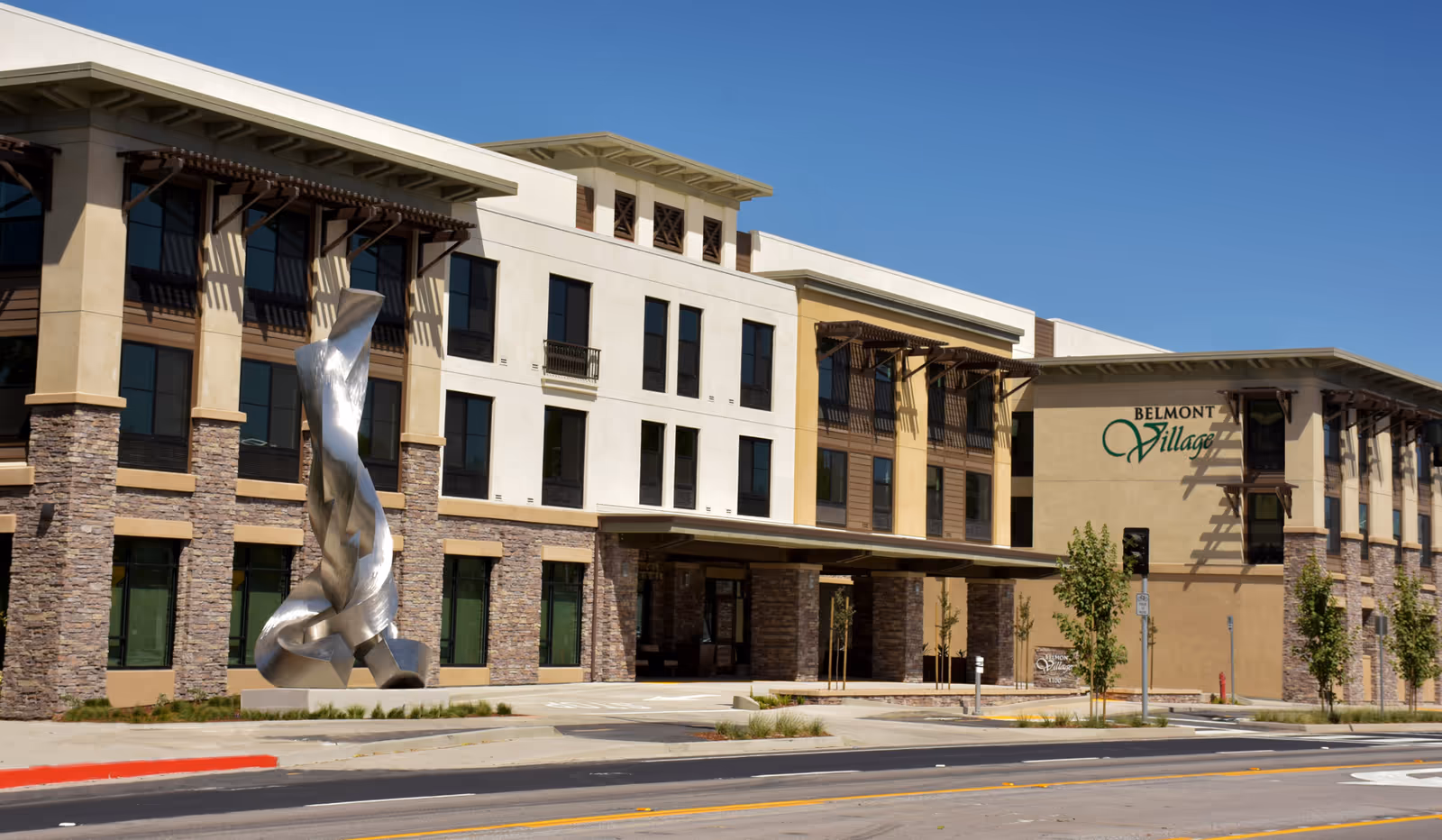 Exterior view of Belmont Village Senior Living Albany building with a modern multi-story design, stone and stucco facade, large windows, and a tall abstract metal sculpture near the entrance under a clear blue sky.