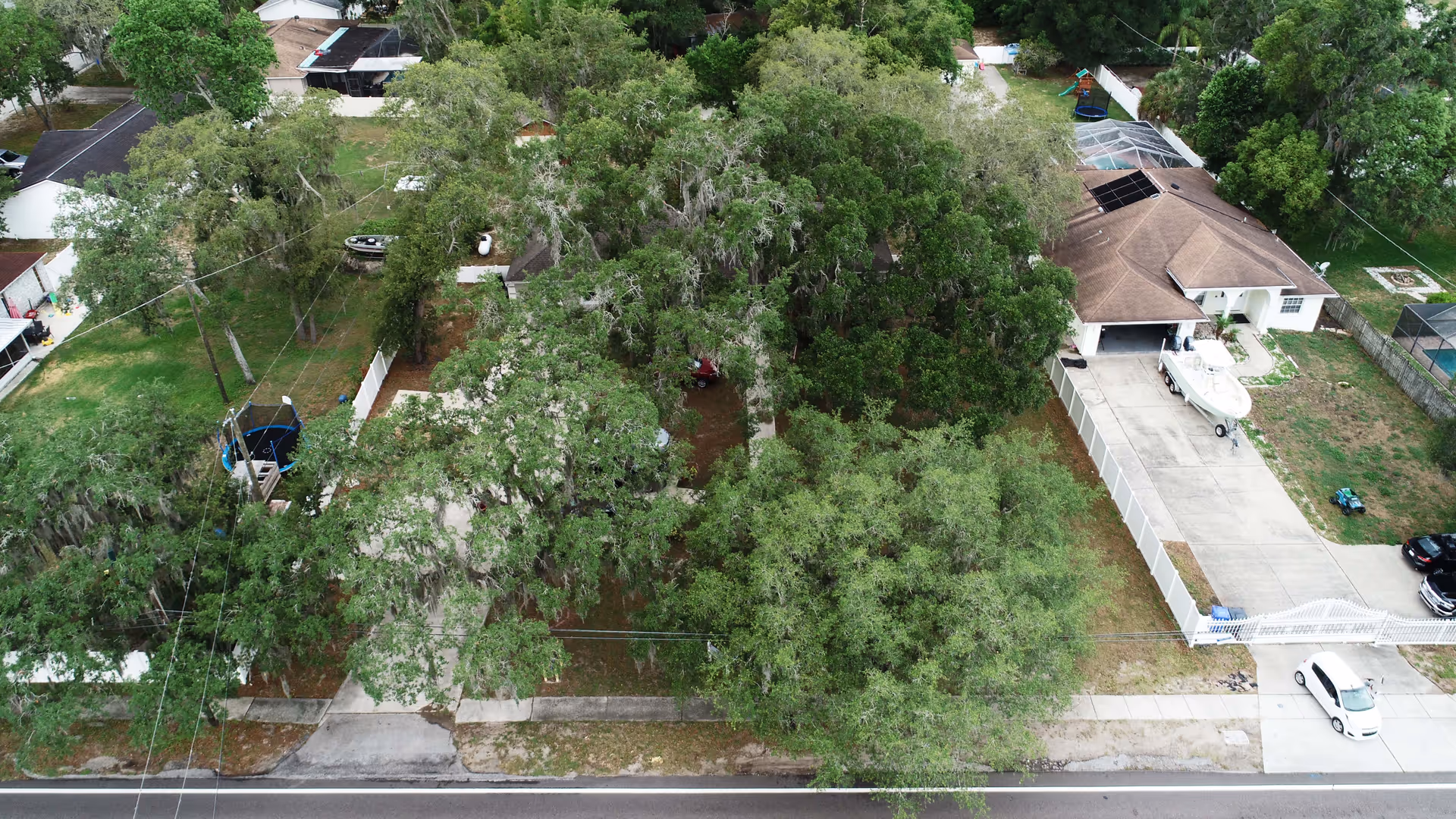 Aerial view of a residential neighborhood showing a house with a large driveway, a boat parked on the driveway, and several trees surrounding the property. The house has a brown roof and is enclosed by a white fence. Nearby houses and green lawns are also visible.