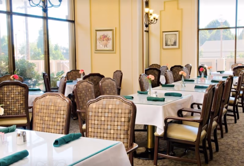 Dining room with multiple tables covered in white tablecloths, each set with green napkins and small flower vases. The room has large windows letting in natural light, framed artwork on the walls, and cushioned chairs with patterned backs.