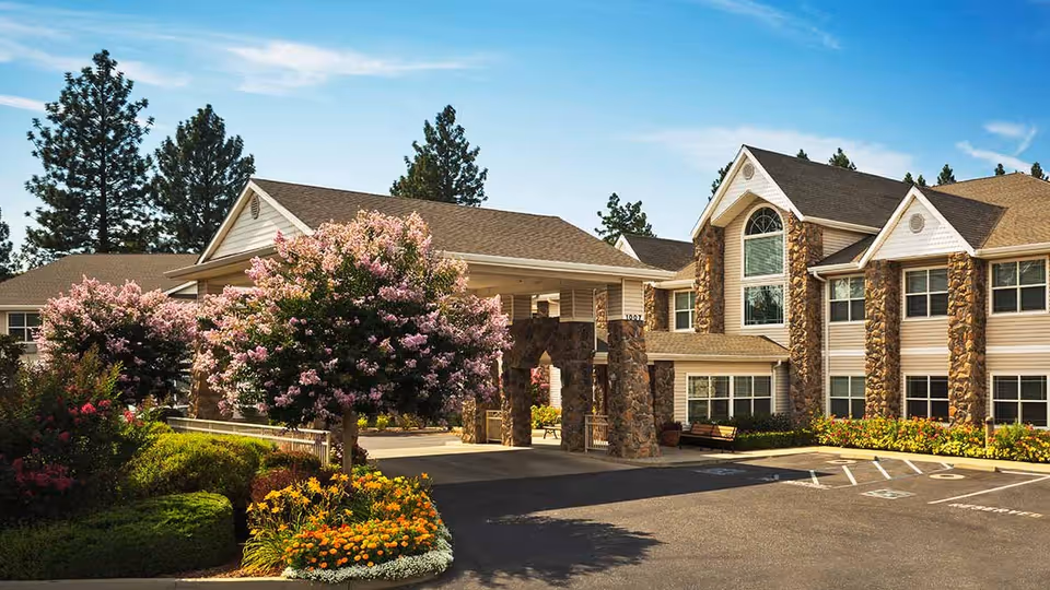 Exterior view of Atria Paradise senior living facility showing a large building with stone pillars, multiple windows, and a covered entrance. The surrounding area features blooming pink and yellow flowers, green shrubs, and tall pine trees under a clear blue sky.