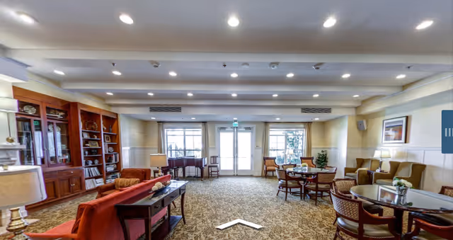 Spacious communal lounge with sofas, armchairs, tables, a bookshelf and a piano facing glass doors.