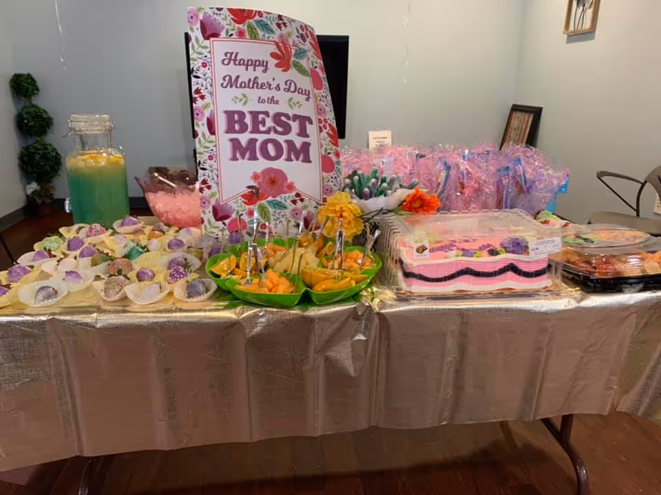 A table set up with various food items including fruit trays, decorated cupcakes, a large pink and white cake, and a pitcher of green and yellow beverage. A sign on the table reads 'Happy Mother's Day to the BEST MOM' with floral decorations. The table is covered with a shiny beige tablecloth and there are chairs and wall decorations in the background.