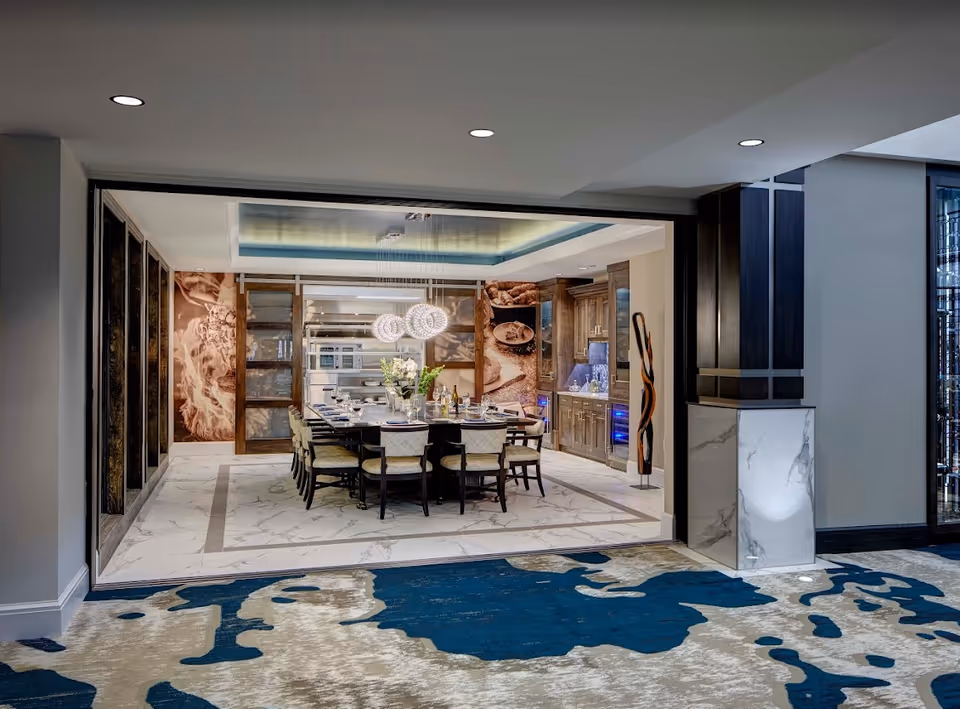 Elegant open dining room with a large rectangular table set for guests, modern pendant lights, and a view into a kitchen area with cabinetry.