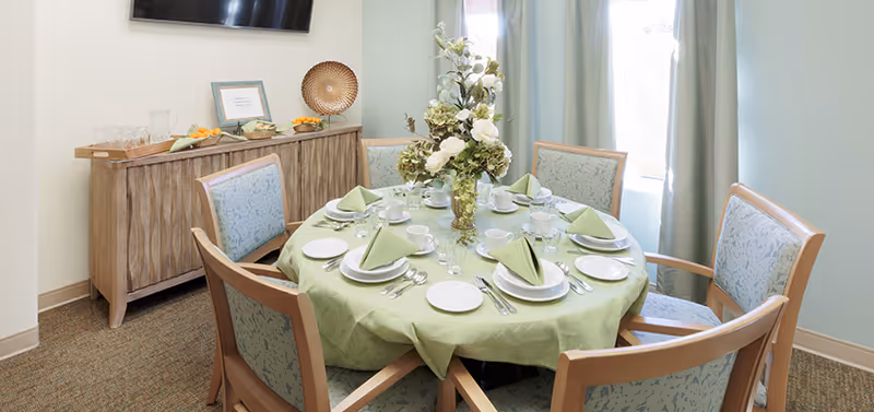 A round dining table set for six with green tablecloth and matching folded napkins, white plates, cups, and silverware. A floral centerpiece is in the middle of the table. Behind the table is a wooden sideboard with glasses, fruit, and decorative items. A flat-screen TV is mounted on the wall above the sideboard. Light green curtains frame a window letting in natural light.