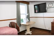 A simple, clean room with a single bed covered in a pink blanket, a small wooden table with a white orchid plant near a window with green curtains, a wall-mounted flat screen TV, a clock, and a calendar on the wall.