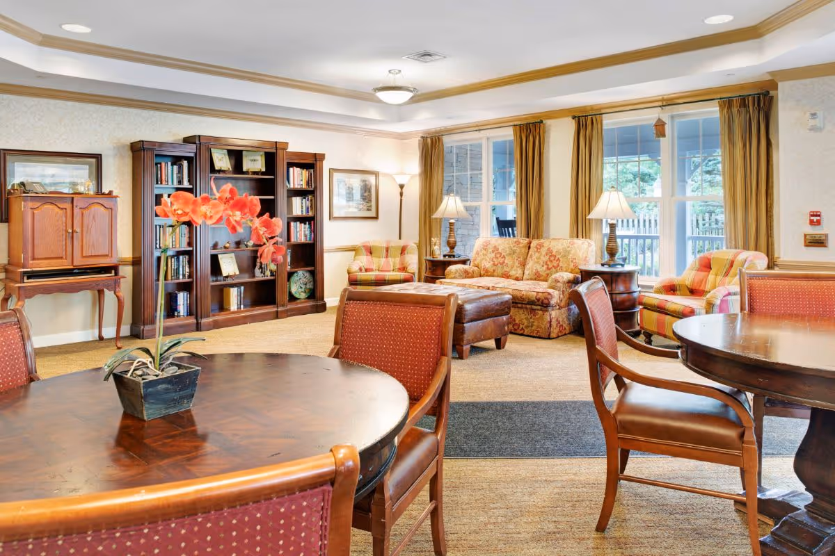 Bright common room with round wooden tables and chairs, a floral sofa and armchairs, a bookcase, and large windows.