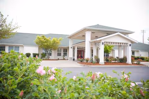 Front view of a single-story memory care facility with a covered entrance and blooming bushes.