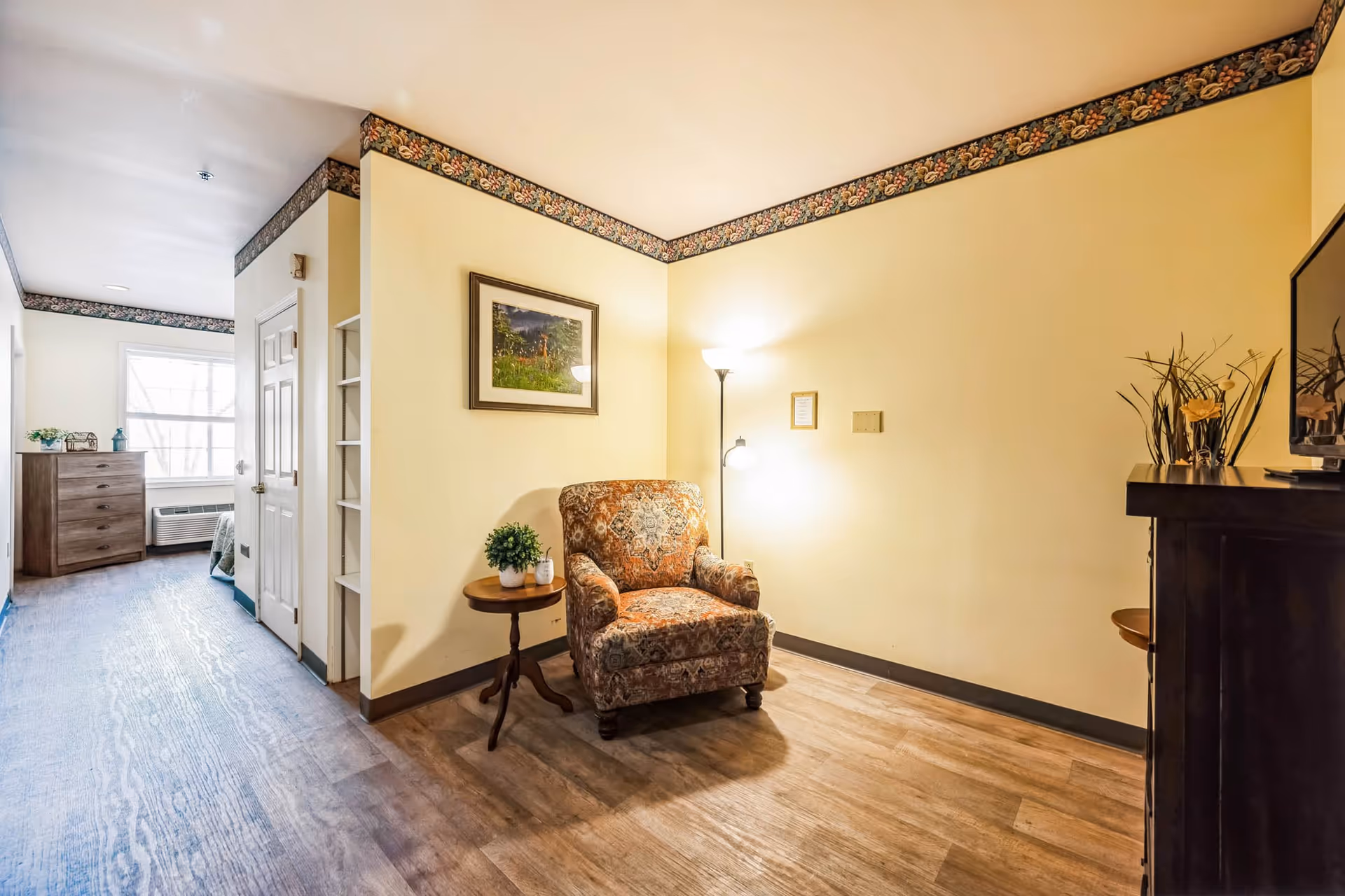 A cozy living area in a senior living facility featuring a patterned armchair next to a small round wooden table with a potted plant and a candle. The walls are painted light yellow with floral wallpaper border near the ceiling. A floor lamp with two lights illuminates the corner. A framed picture hangs on the wall above the chair. To the left, a hallway leads to a bedroom with a dresser and a window letting in natural light. On the right side, a TV sits on a dark wooden stand with decorative plants.