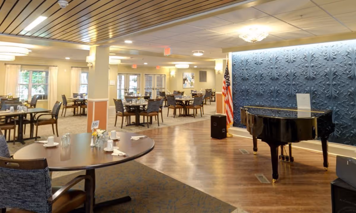 A spacious dining room in a senior living facility with multiple tables and chairs arranged neatly. The tables are set with cups, glasses, and napkins. To the right, there is a black grand piano placed on a wooden floor area with a decorative blue textured wall behind it and an American flag standing next to it. The room is well-lit with ceiling lights and has large windows with curtains allowing natural light to enter.