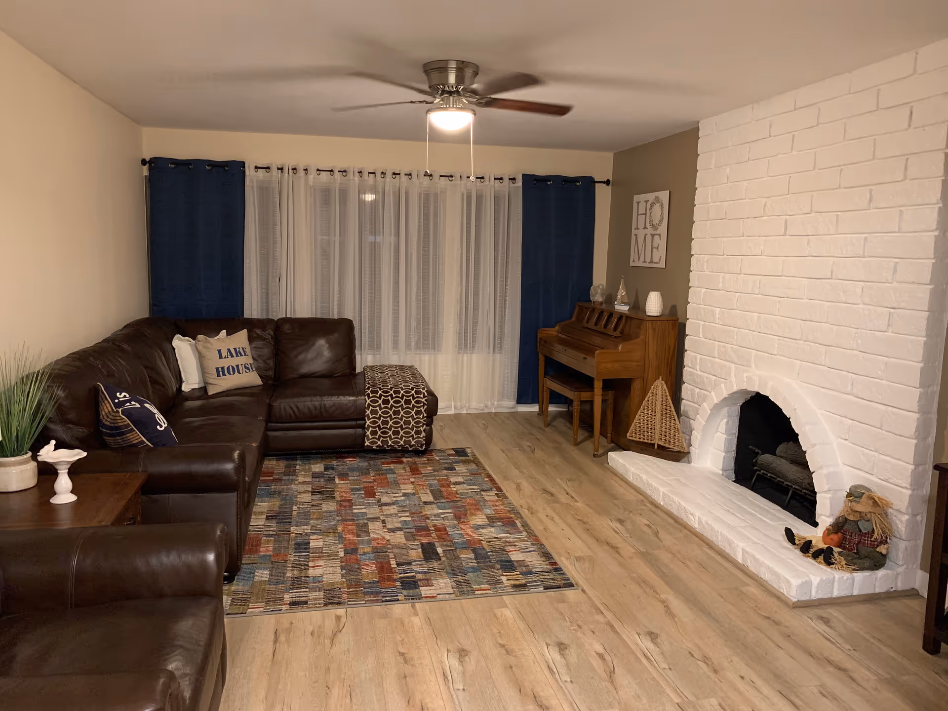 A cozy living room with a large brown leather sectional sofa adorned with decorative pillows, a colorful patterned rug on a wooden floor, a white brick fireplace with a small scarecrow decoration, a wooden piano against the wall, and blue and white curtains covering the windows. A ceiling fan with a light is mounted on the ceiling.