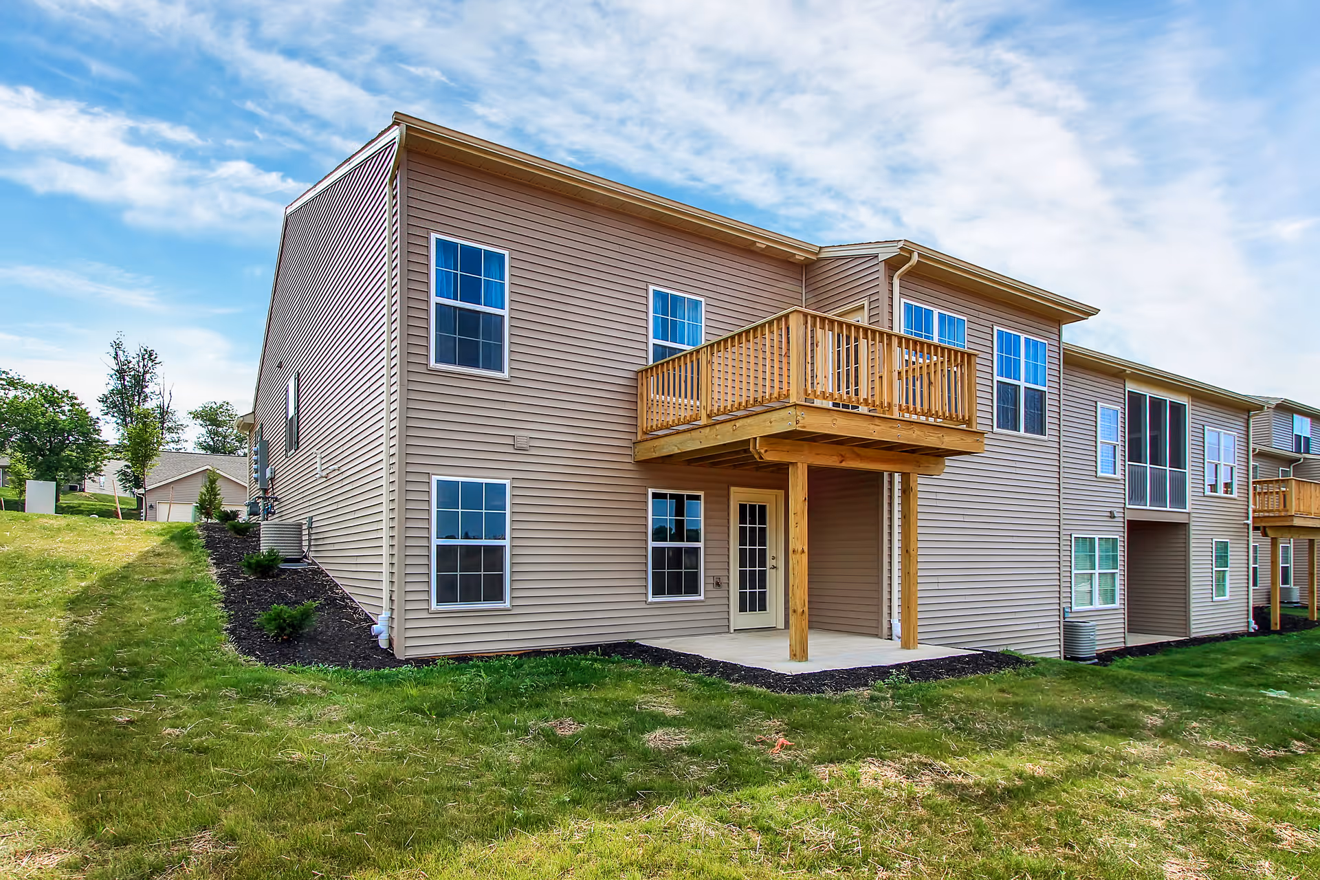 Exterior view of a two-story residential building with beige siding, multiple windows, and a wooden balcony on the upper floor. The building is surrounded by a grassy lawn and a partly cloudy blue sky is visible above.