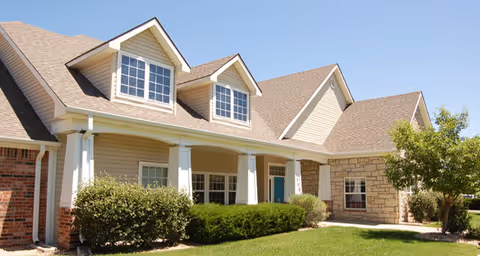 Exterior view of a residential-style building with beige siding, brick and stone accents, white columns supporting a covered porch, and a well-maintained lawn with bushes and a tree under a clear blue sky.