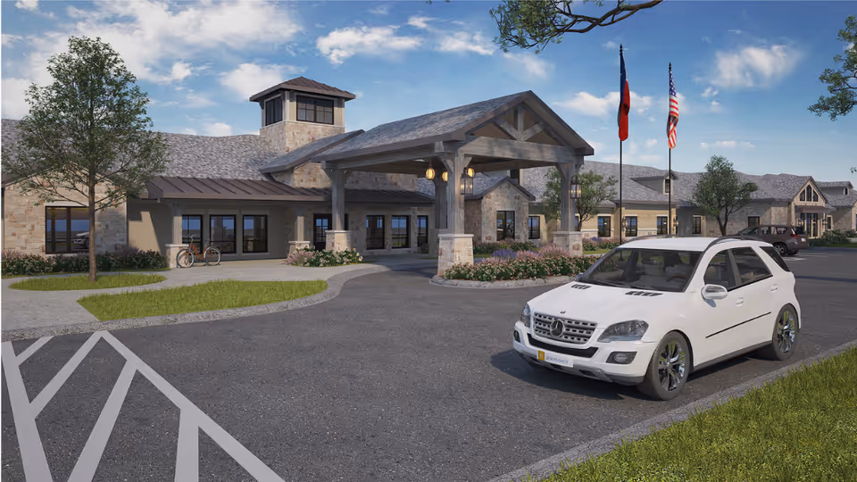 Exterior view of Celebration Senior Living of Denison, showing a large stone building with a covered entrance supported by wooden beams. There are two flagpoles with flags, a white SUV parked in front, trees, and a clear blue sky with some clouds.