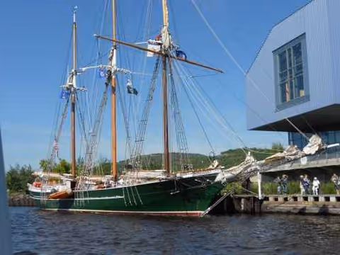 Tall-masted sailing ship docked beside a modern building by calm water