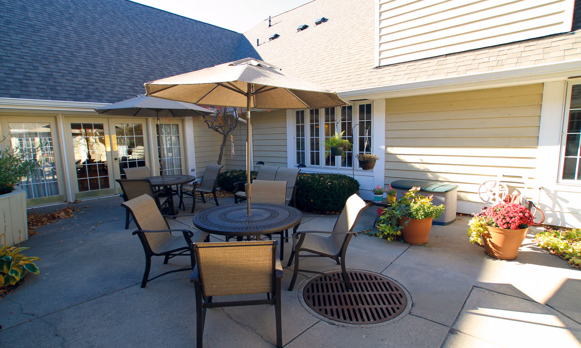 Outdoor patio area with round tables and chairs under large umbrellas, surrounded by potted plants and bushes, adjacent to a beige building with multiple windows and glass doors.