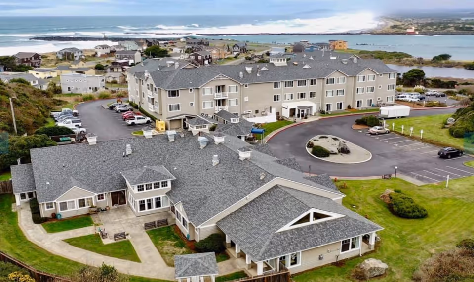 Aerial view of a senior living community's beige multi-story buildings, parking lot and landscaped grounds with the ocean in the background.