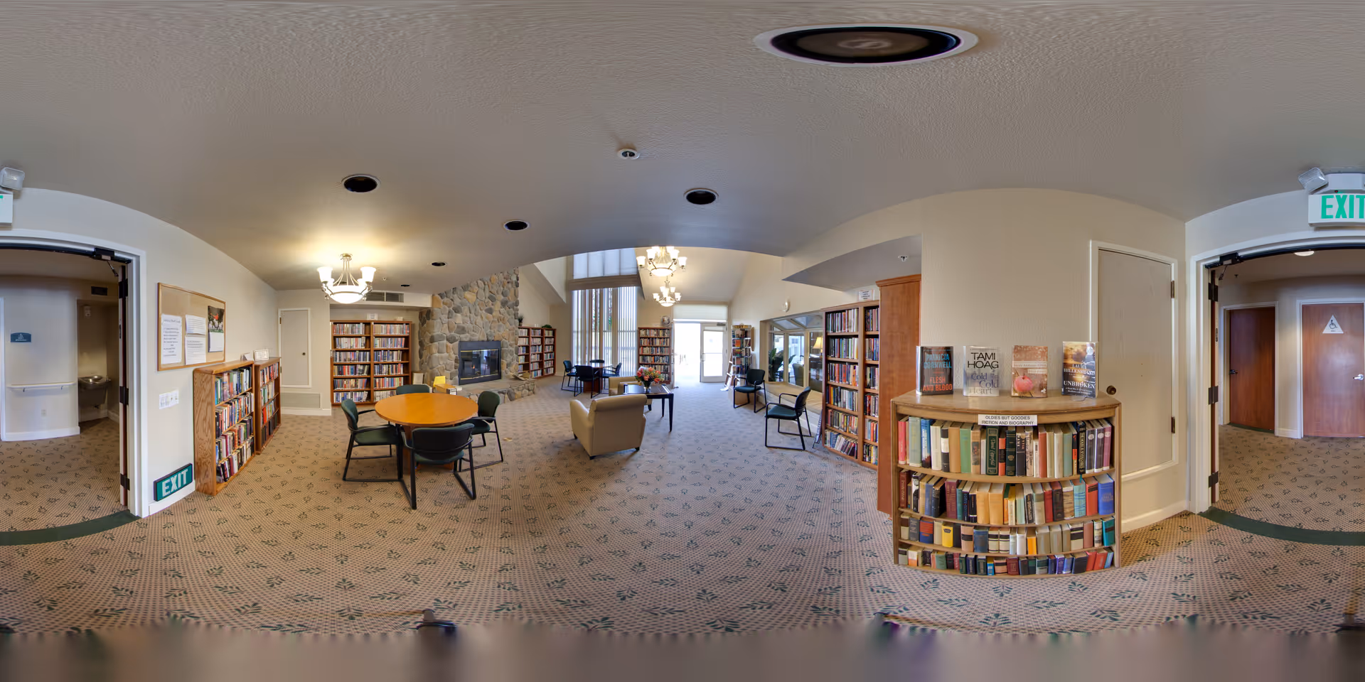 A spacious senior living facility common area with carpeted floors and beige walls. The room features multiple bookshelves filled with books, a round wooden table with chairs, a stone fireplace, and comfortable seating including armchairs. Large windows allow natural light to fill the space. There are exit signs above doorways on either side of the room.