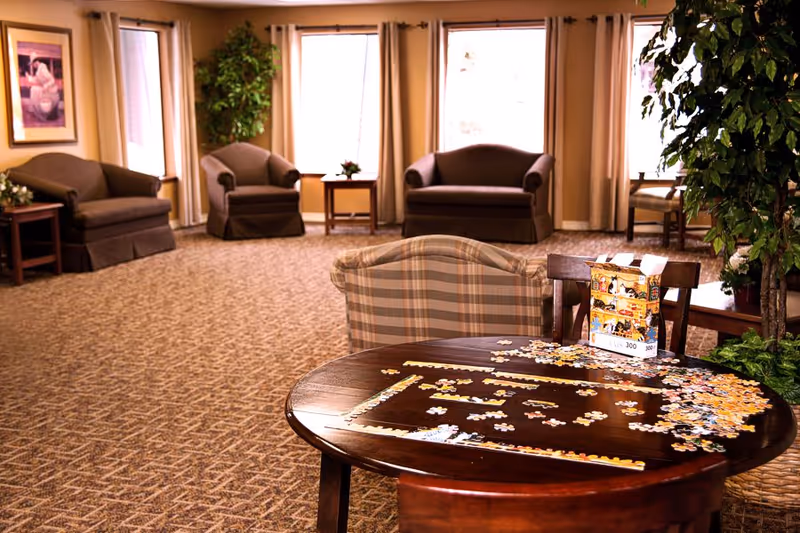 A spacious living room area with several armchairs and loveseats arranged around the perimeter. In the foreground, a round wooden table holds a partially completed jigsaw puzzle with scattered puzzle pieces and the puzzle box. Large windows with curtains allow natural light to fill the room, and there are potted plants adding greenery to the space.