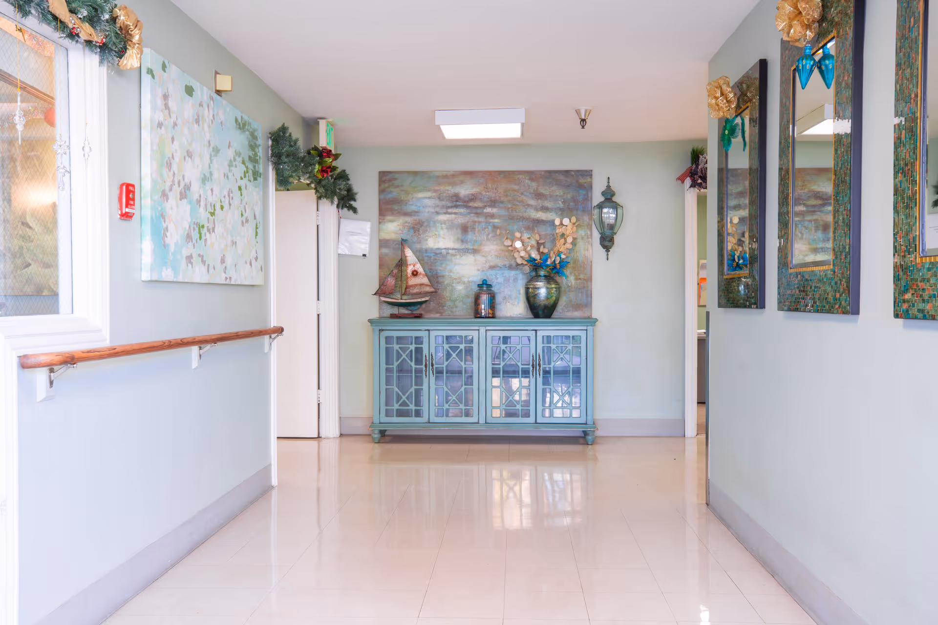 A clean, well-lit hallway in a senior living facility with light blue walls, a wooden handrail on the left side, and decorative artwork including a painting, a model sailboat, and a vase with flowers on a blue cabinet at the end of the hallway. There are also three framed mirrors on the right wall and holiday decorations near the ceiling.