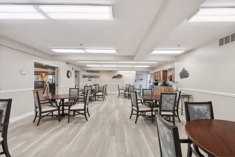 A spacious dining room with multiple round wooden tables and cushioned chairs arranged neatly. The room has light-colored walls and flooring, with bright overhead fluorescent lighting. On the far wall, there is a sign that reads 'ASHWOOD' along with some framed pictures and a decorative wall piece. A kitchen area with wooden cabinets and appliances is visible on the right side of the room.