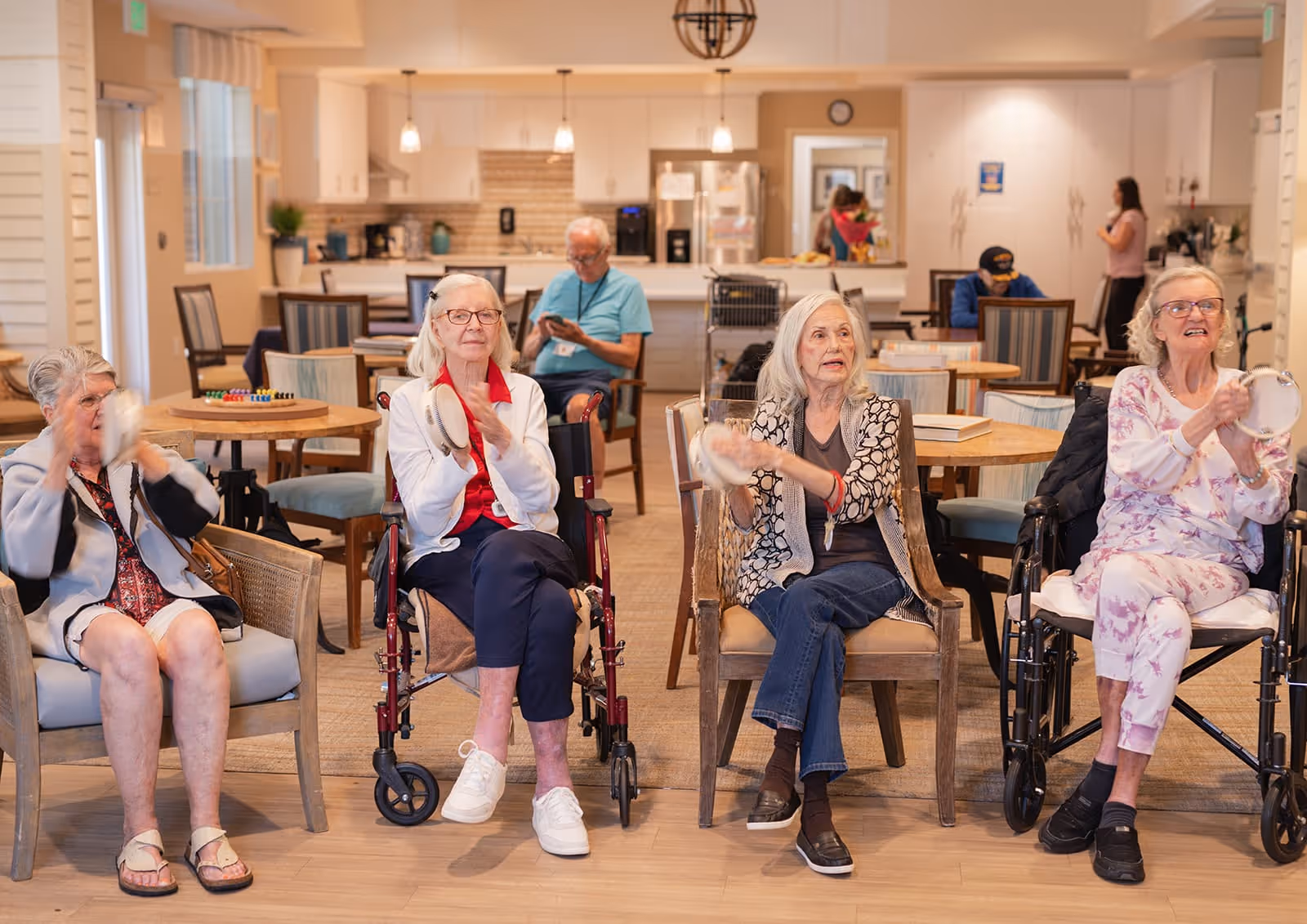 Four elderly women sitting in chairs and wheelchairs in a common room, playing tambourines. In the background, there are tables, chairs, and a kitchen area with a man and a woman engaged in different activities.
